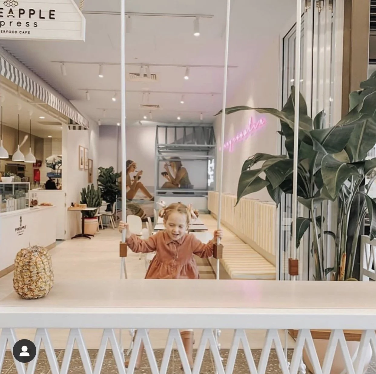 A young girl with pigtails wearing a brown dress, playing on an indoor swing in a cafe. The cafe has modern decor, with plants, a neon sign that says 'gorgeous,' and customers sitting at tables, including two women near the window.