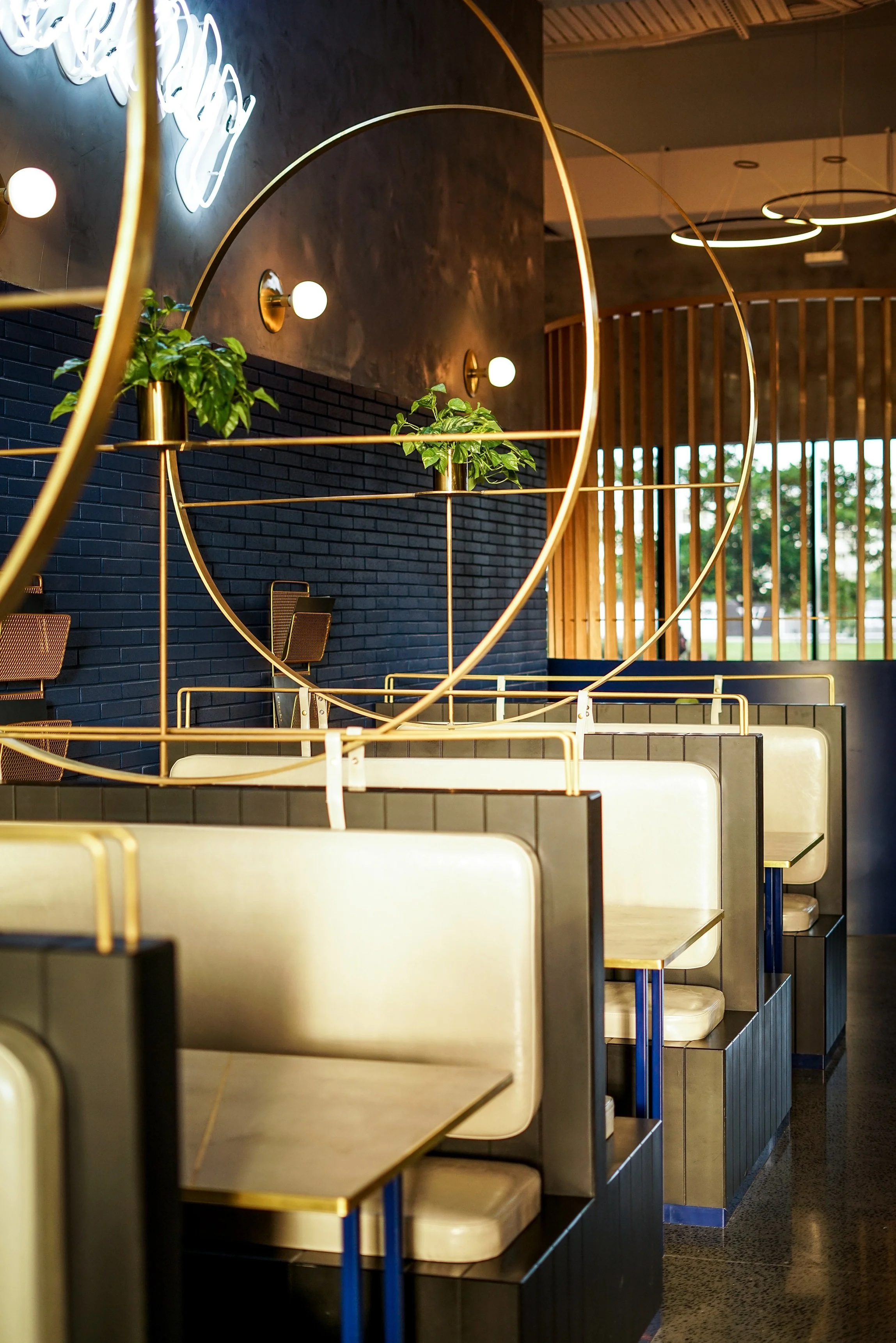 Empty modern booth seating at a restaurant with black brick wall, gold accents, potted plants, and large windows in the background.