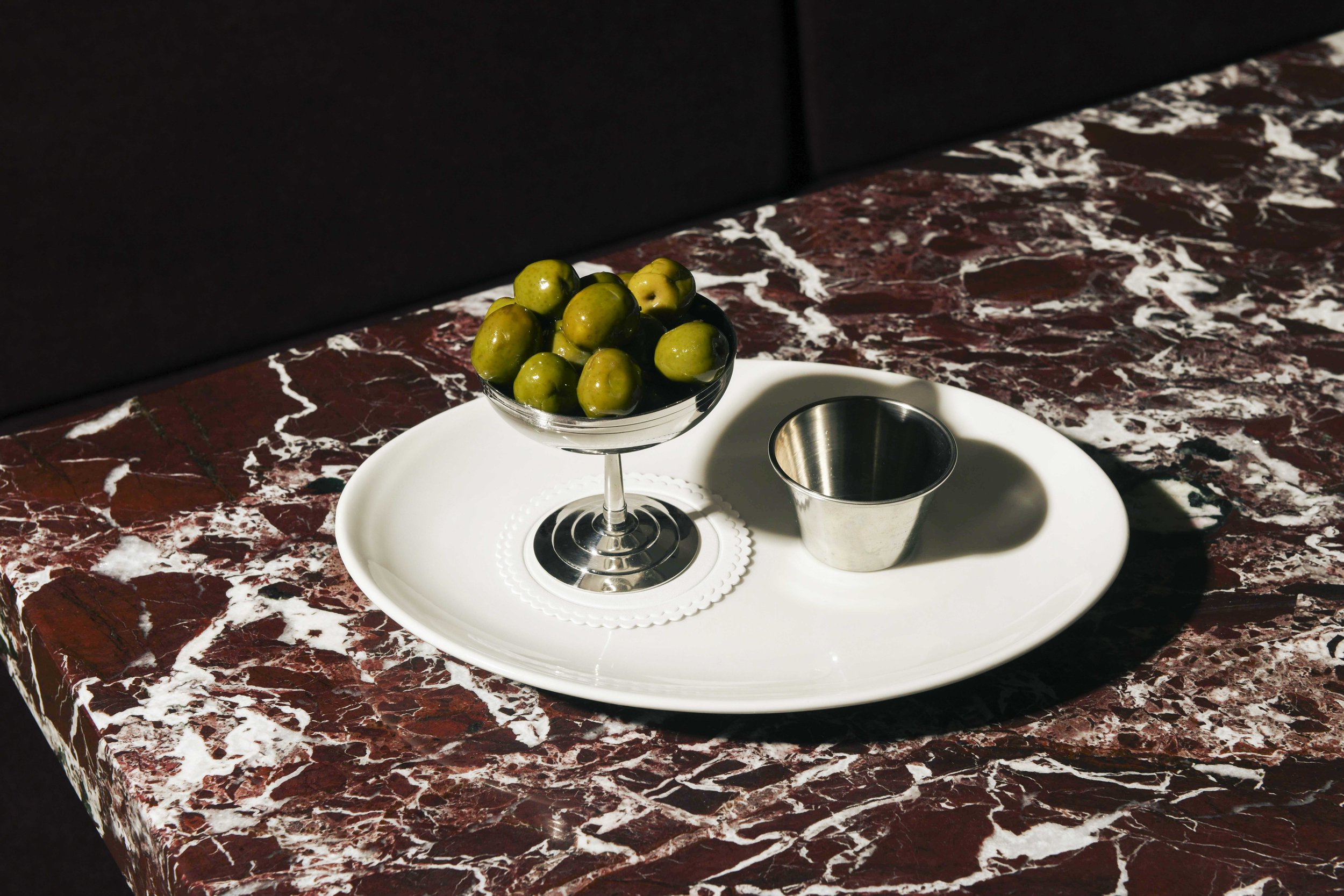 A white oval tray holding a glass bowl of small green apples and a metal cup on a dark red marble surface.