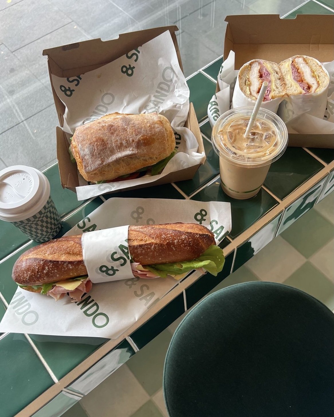 Three sandwiches on a tiled counter, each wrapped in paper with "Sando" printed on it. One sandwich is in a ciabatta roll, another in a baguette, and the third is halved, showing layered fillings. Beside them are cups of iced and hot coffee.