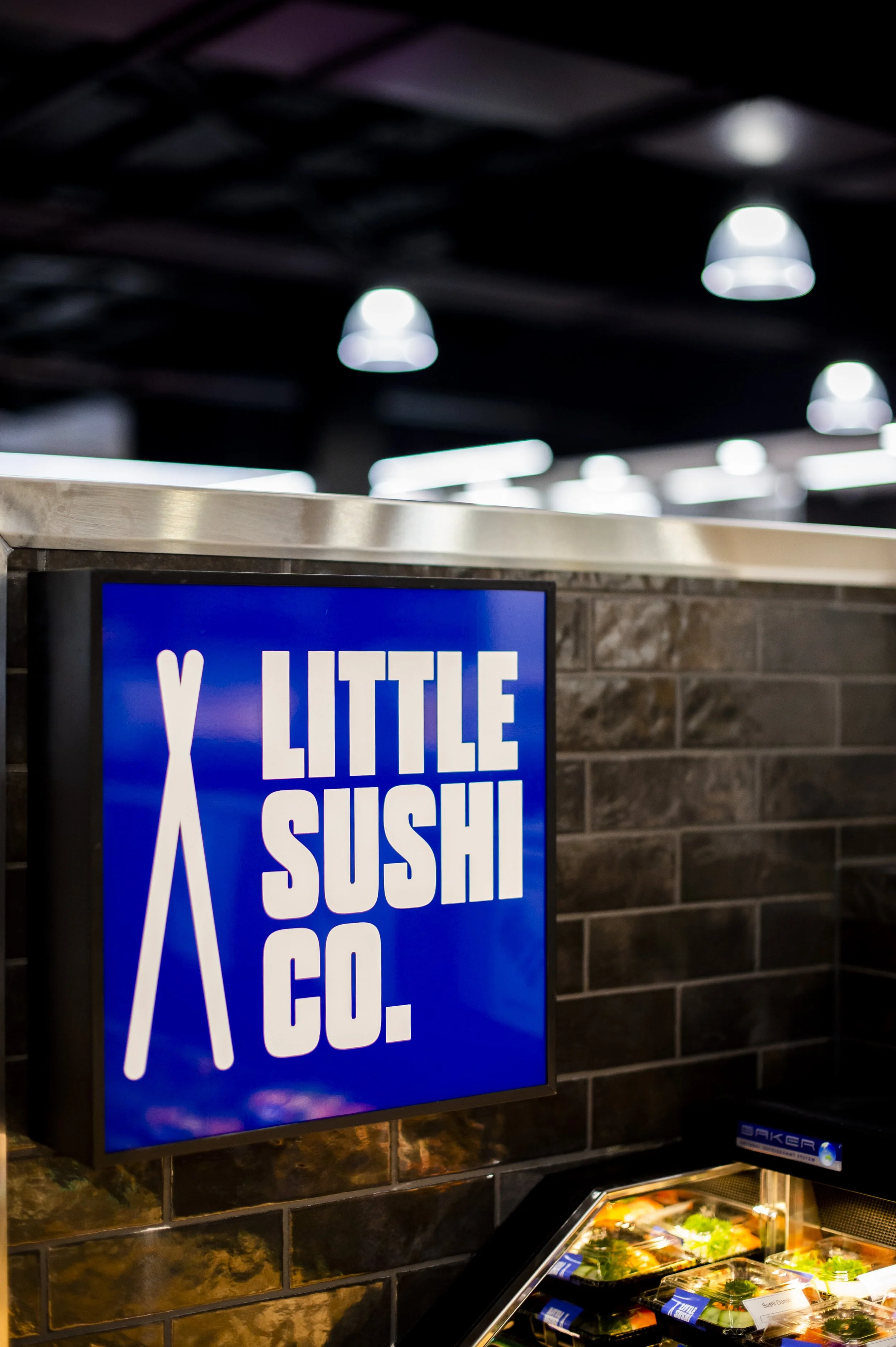 Sign for Little Sushi Co., a sushi restaurant, with a display case of sushi containers below.