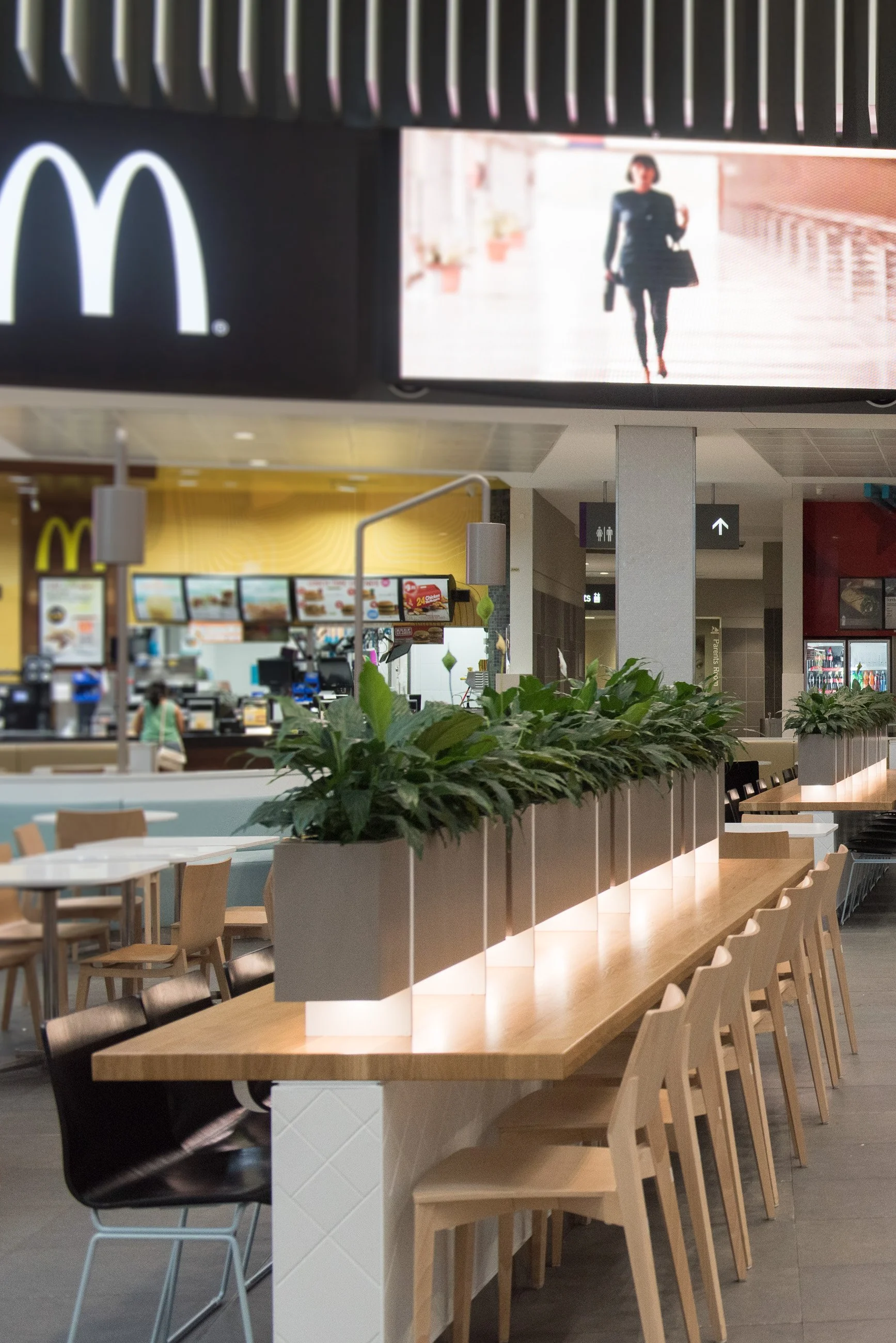 Interior of a McDonald's restaurant with a long wooden table, beige chairs, and a row of green plants in a gray planter dividing the seating area. The counter and menu boards are visible in the background, and a large digital advertising screen displ