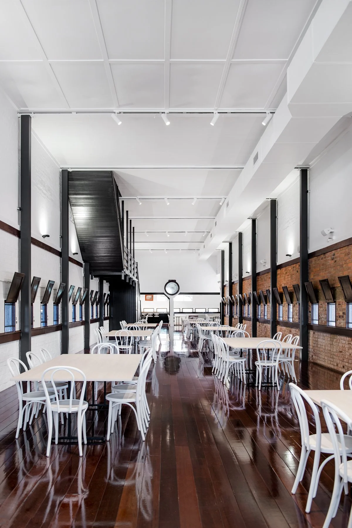Empty modern cafeteria with white chairs, light-colored tables, dark wooden floors, brick and white walls, black windows, and overhead lights.