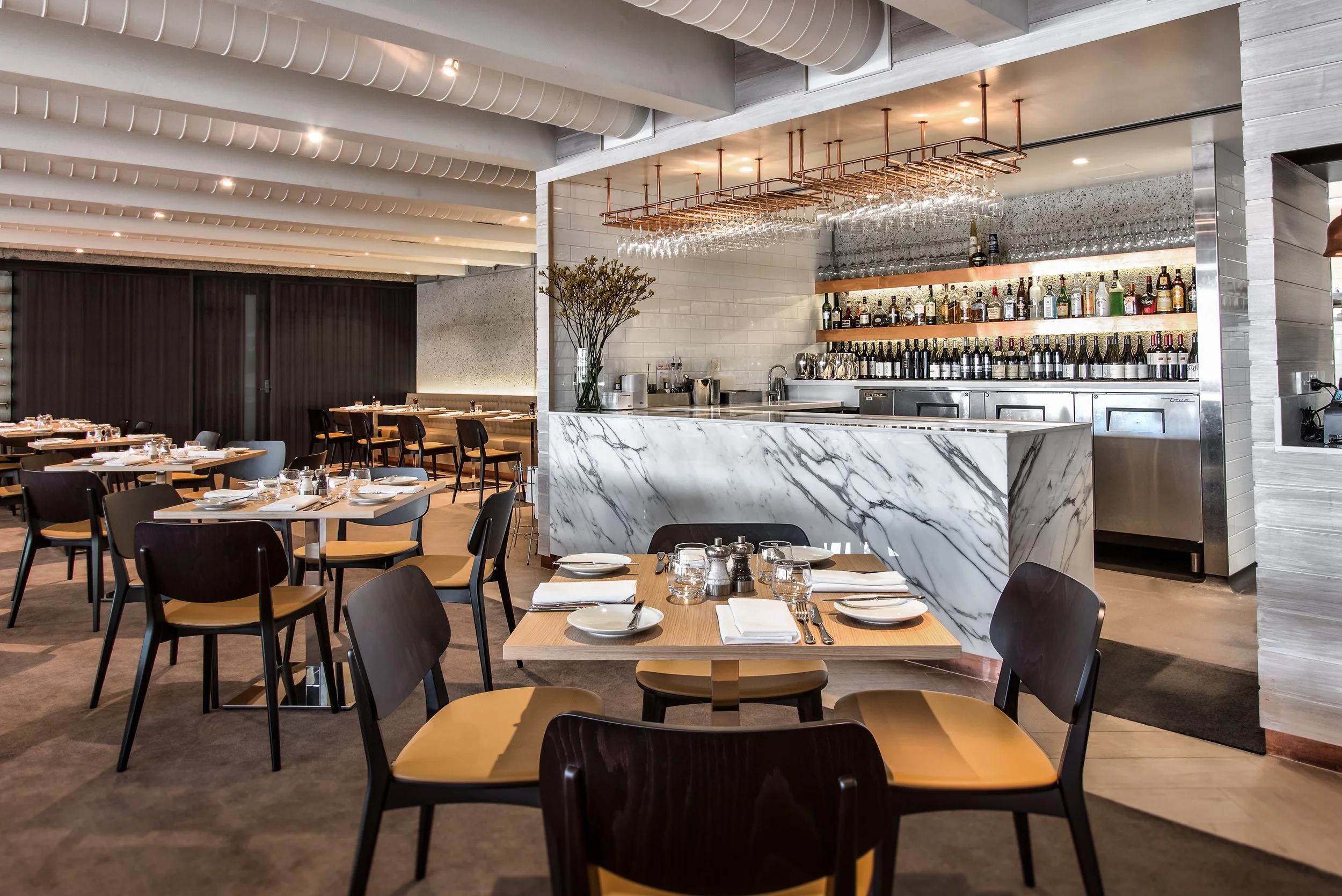 Interior of a modern restaurant with tables set for dining, a marble bar counter, and a well-stocked bar with bottles and glasses on shelves.