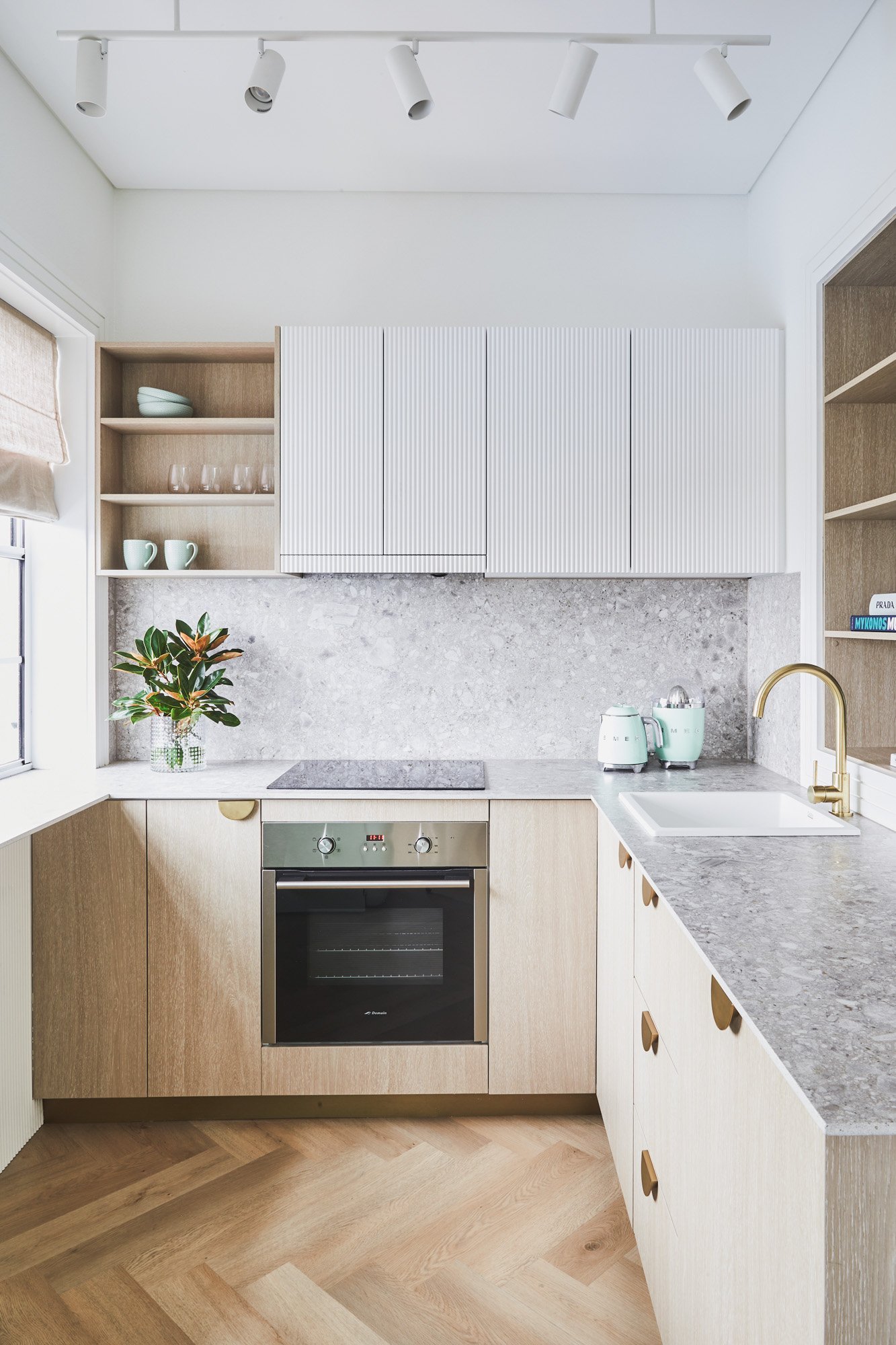 Modern kitchen with wooden cabinets, gray countertops, built-in oven, and open shelving. Features a brass faucet, white sink, and decorative plants.