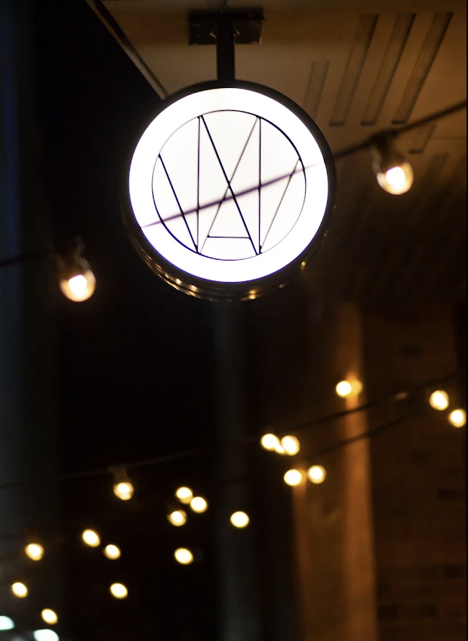 Illuminated round draft beer sign with a black outline and geometric design, hanging from the ceiling in a dimly lit space with warm-colored string lights.