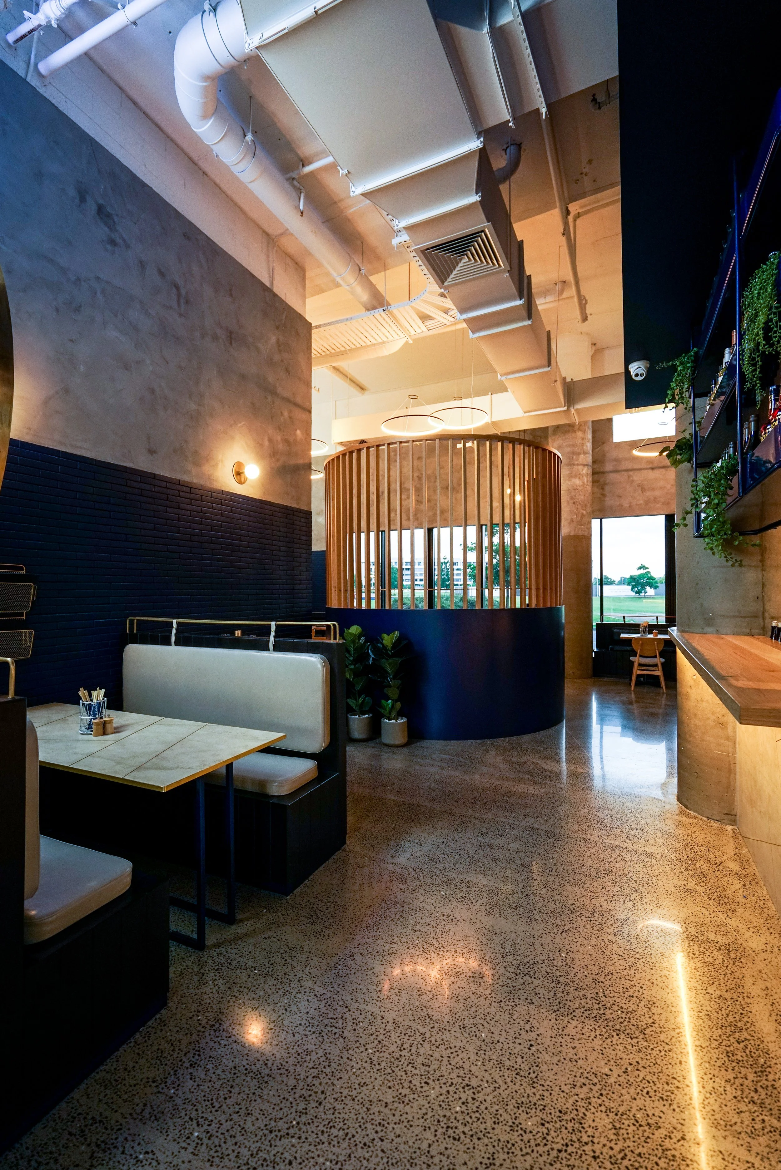 Interior of a modern restaurant with high ceilings, concrete walls, a booth seating area, indoor plants, and large windows showing a view outside.