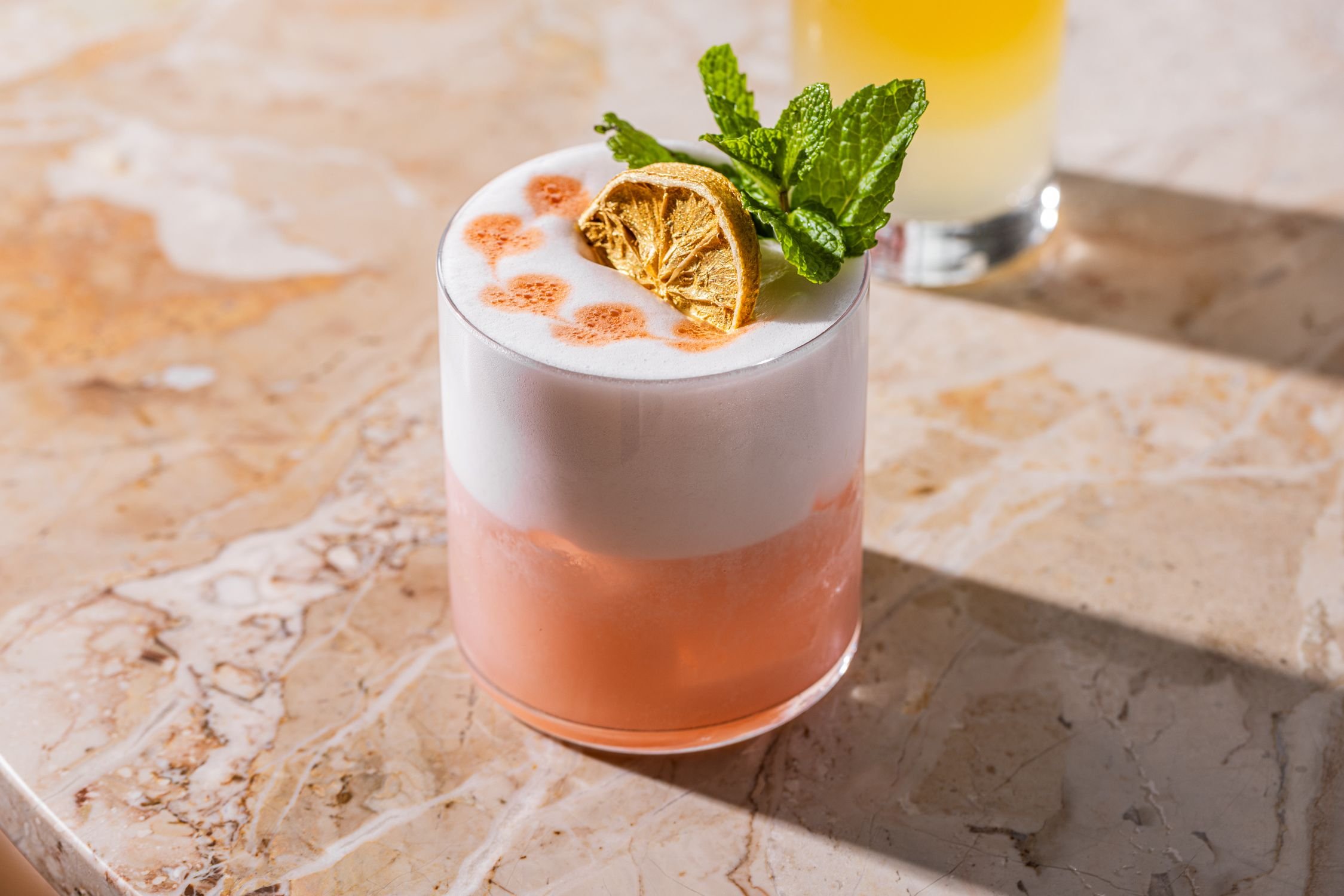 Pink cocktail with foam, garnished with dried citrus slice and mint on marble surface.
