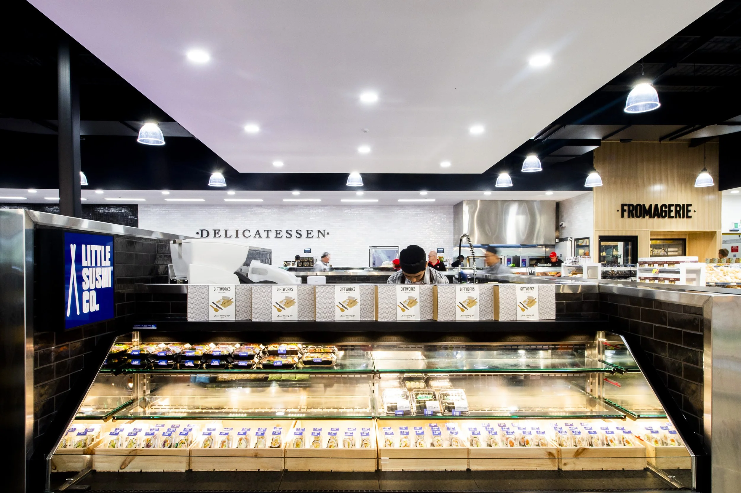 Inside a grocery store with a deli section, sushi counter, and cheese display, illuminated by ceiling lights.