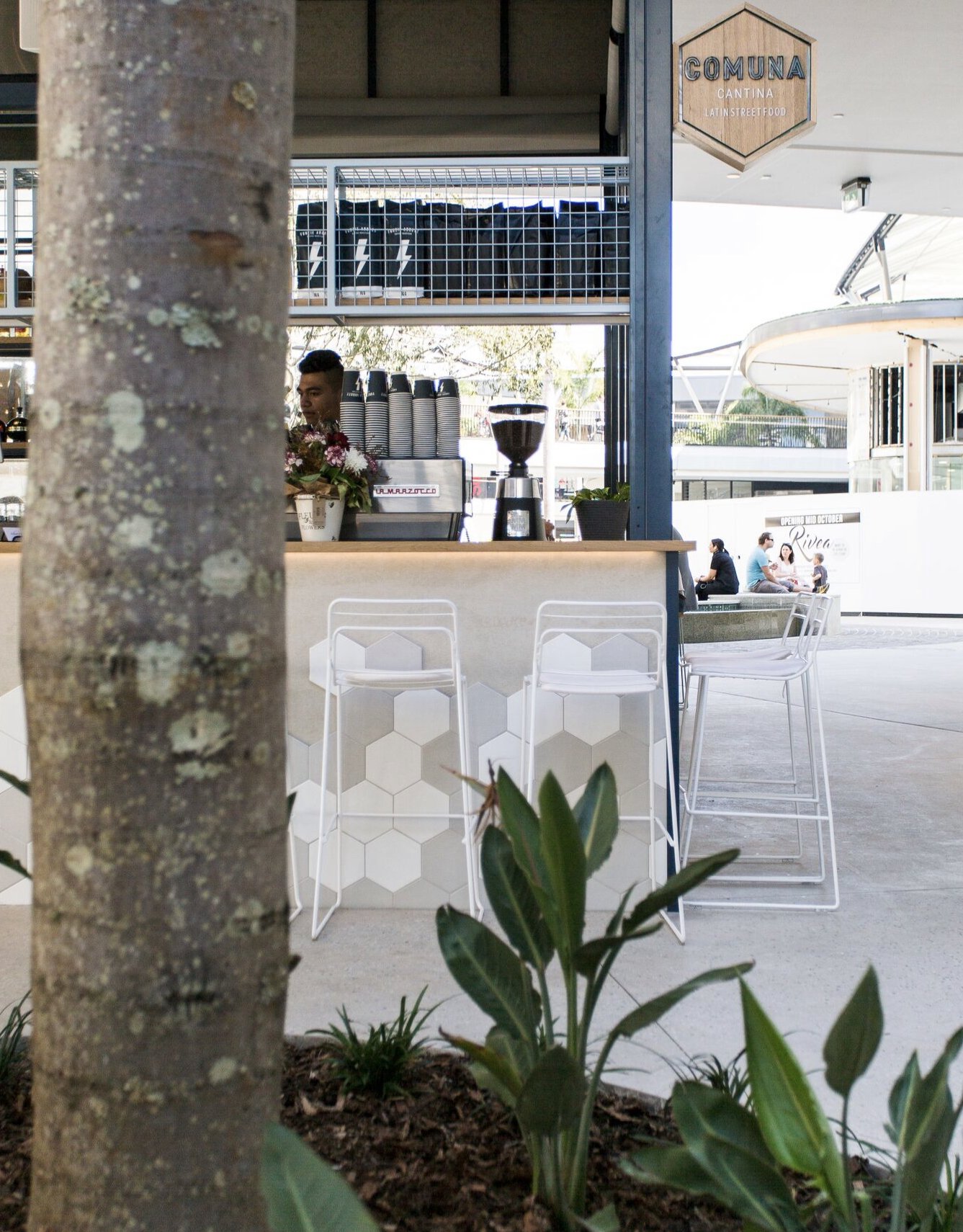 Outdoor scene of a cafe with white barstools, a wooden counter, and a coffee machine. In the background, there are people sitting by a pool and a sign that reads 'COMUNA CANTINA LATIN STREET FOOD.'