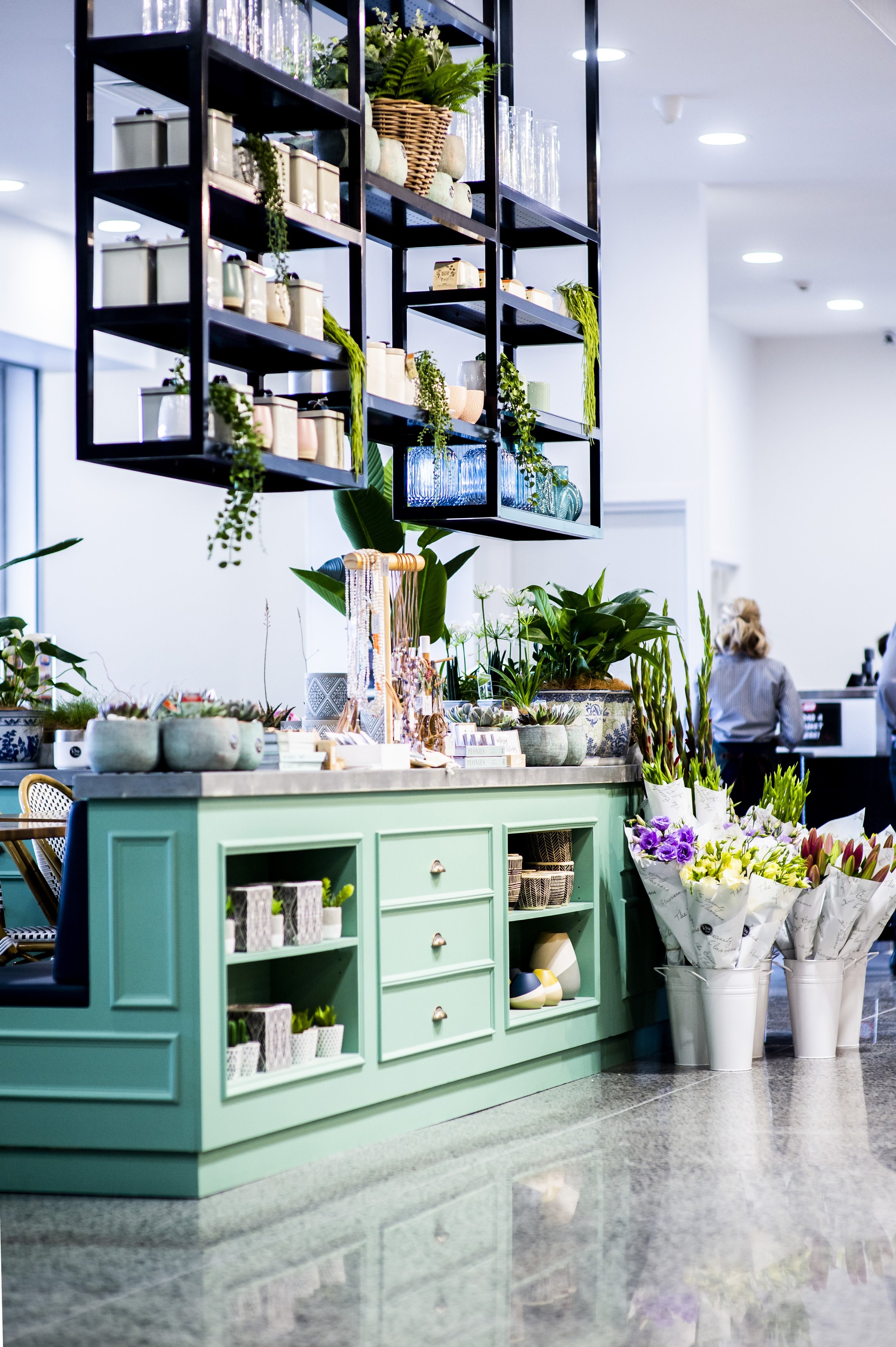 Interior of a modern flower and gift shop with a mint green cabinet, potted plants, artificial flowers, and decorative items on display, with a black metal shelving unit overhead and a woman working in the background.