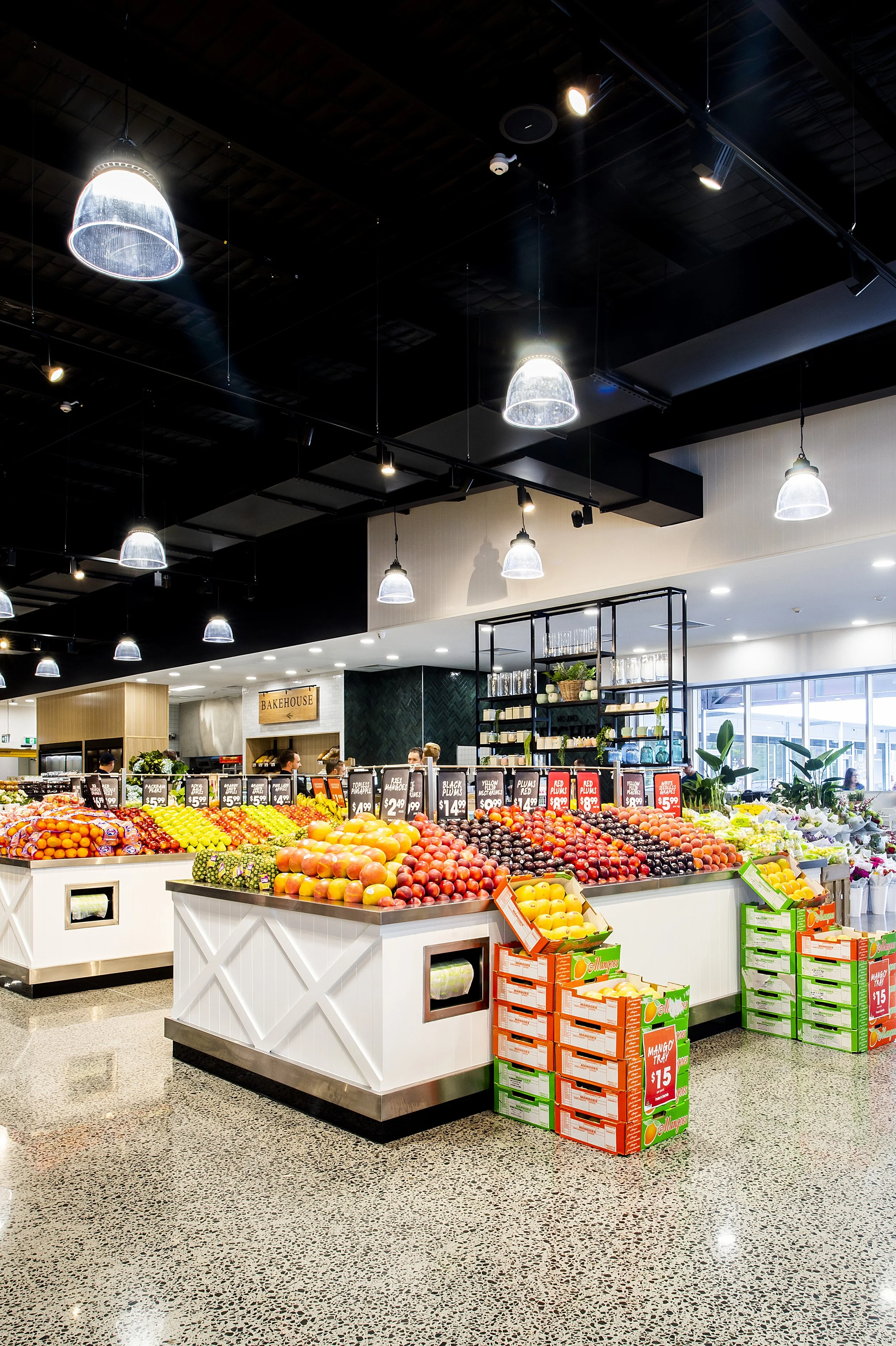 Fruit display in a grocery store with apples, oranges, grapes, and boxes of mangoes under bright lighting.