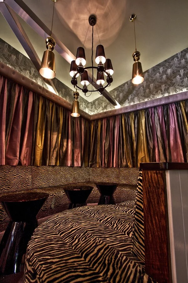 Intimate restaurant or lounge corner with zebra-patterned seating, dark wooden tables, gold and purple curtains, and a black chandelier with multiple lights hanging from the ceiling.