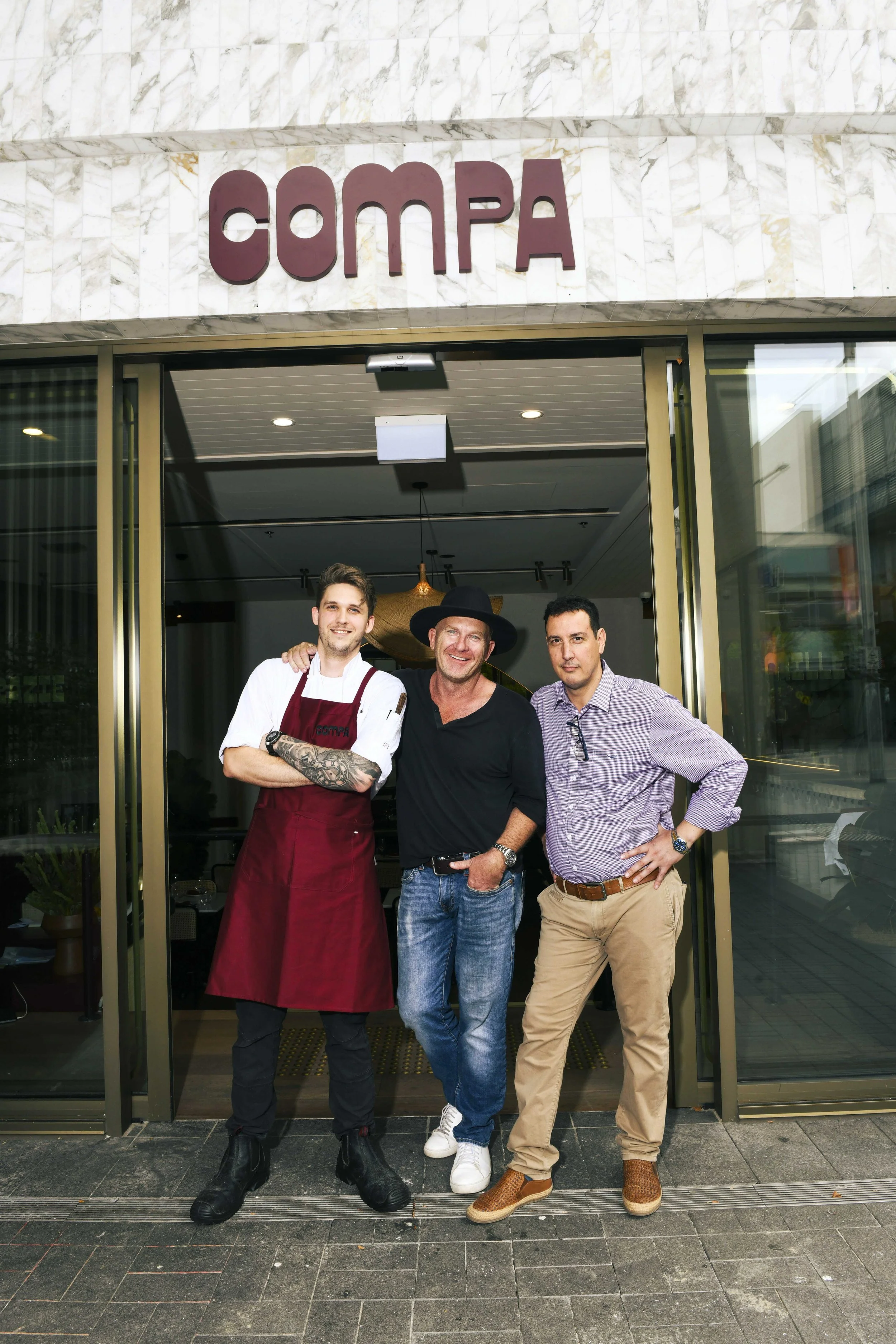 Three men standing in front of the entrance to a restaurant called COMPA, smiling at the camera.