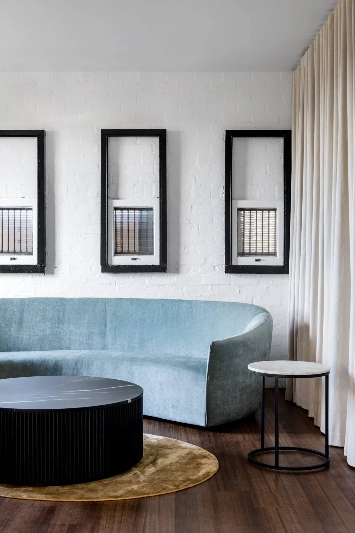Modern living room with light blue curved sofa, round black coffee table, small white marble side table, white brick wall with three rectangular wall art pieces, beige curtains, and hardwood floors.