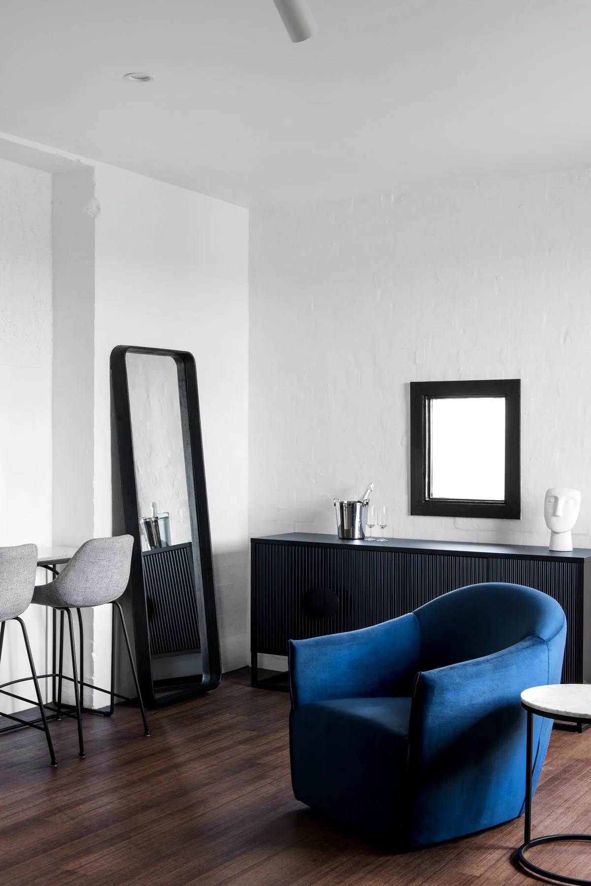 A modern living space featuring a blue velvet armchair, a small side table, a black cabinet with a silver ice bucket and glasses, a large black-framed mirror leaning against the wall, a grey bar stool with a light grey cushion, a small window, and a 