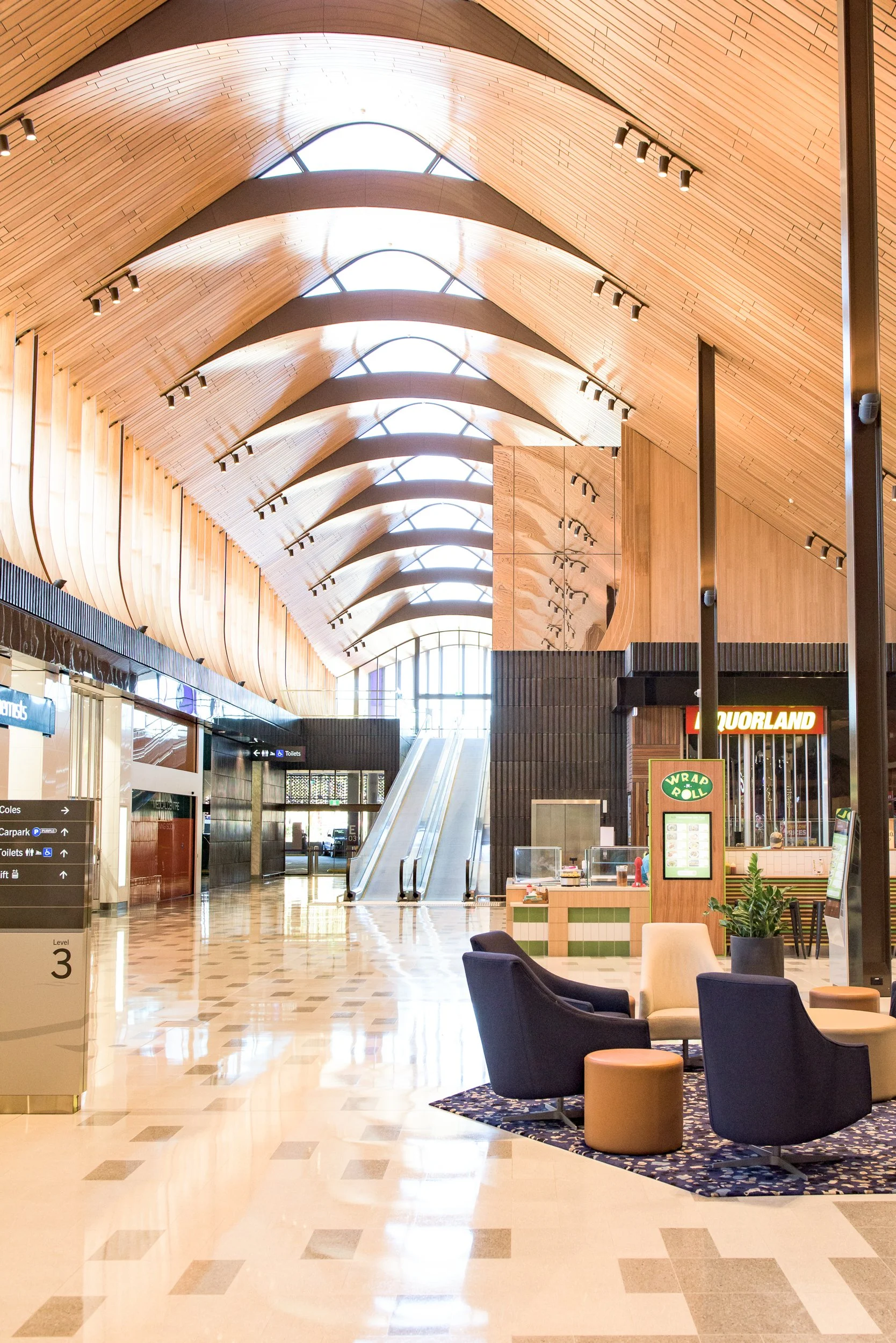 Bright, spacious airport terminal with high, wooden arched ceiling, escalators at the center, and seating area with chairs and a small rug.