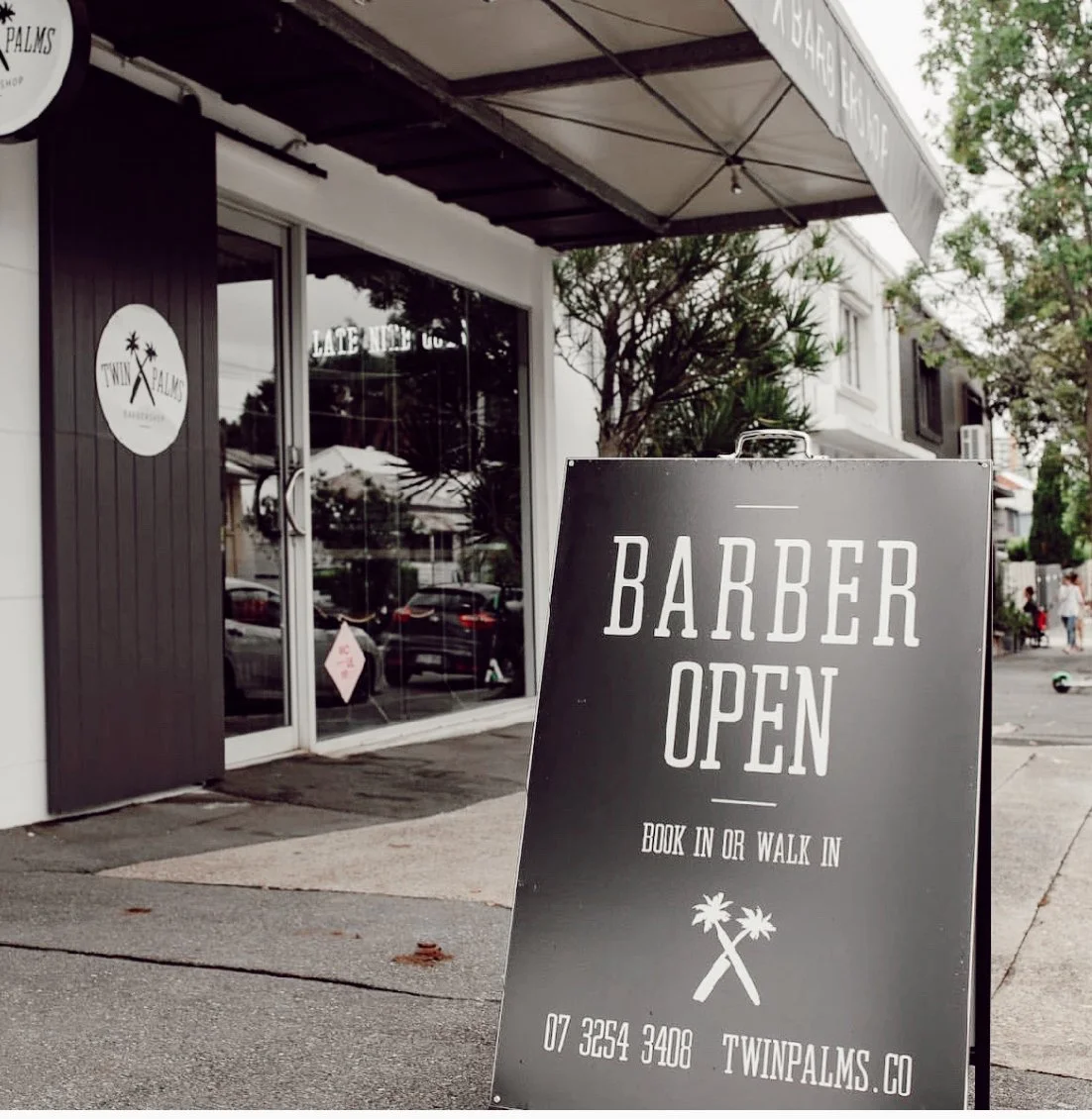 A sidewalk sign outside a barber shop indicating that the barber is open for appointments or walk-ins, with the shop's contact information and website, near a storefront with windows and an awning.