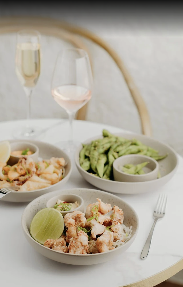 A dining table with bowls of fried seafood, avocado slices, and dipping sauces, with two glasses of white and rosé wine.