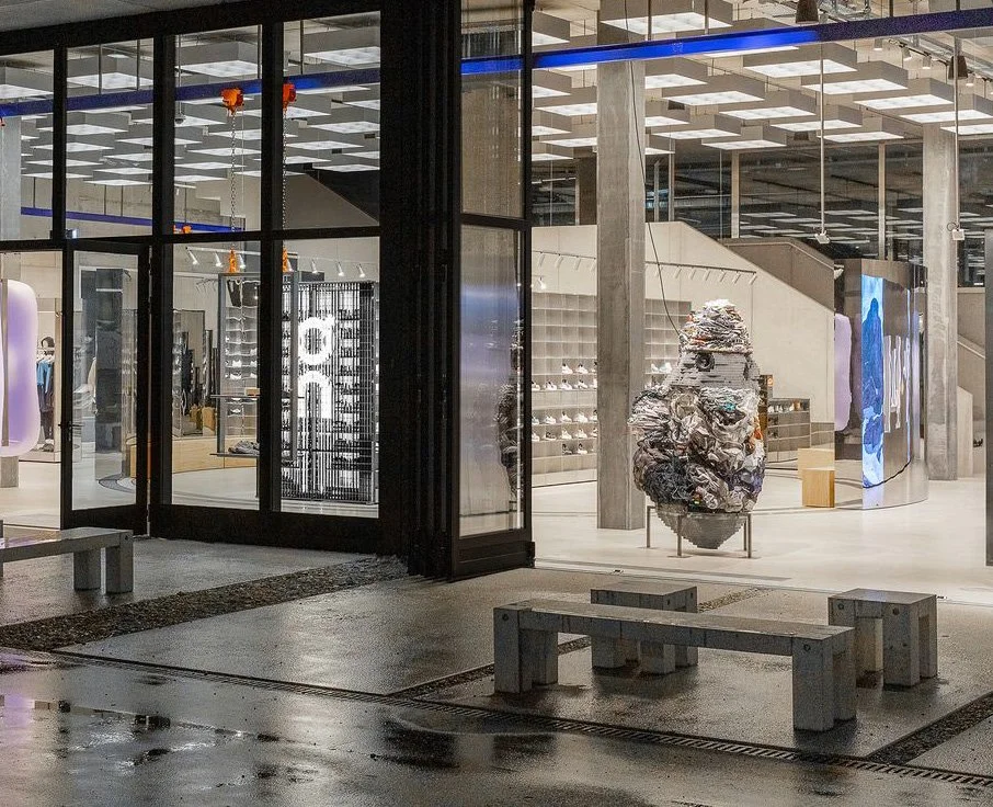 Illuminated On Labs flagship in Zurich viewed through full-height glazing, with concrete seating and reflections on the wet pavement.