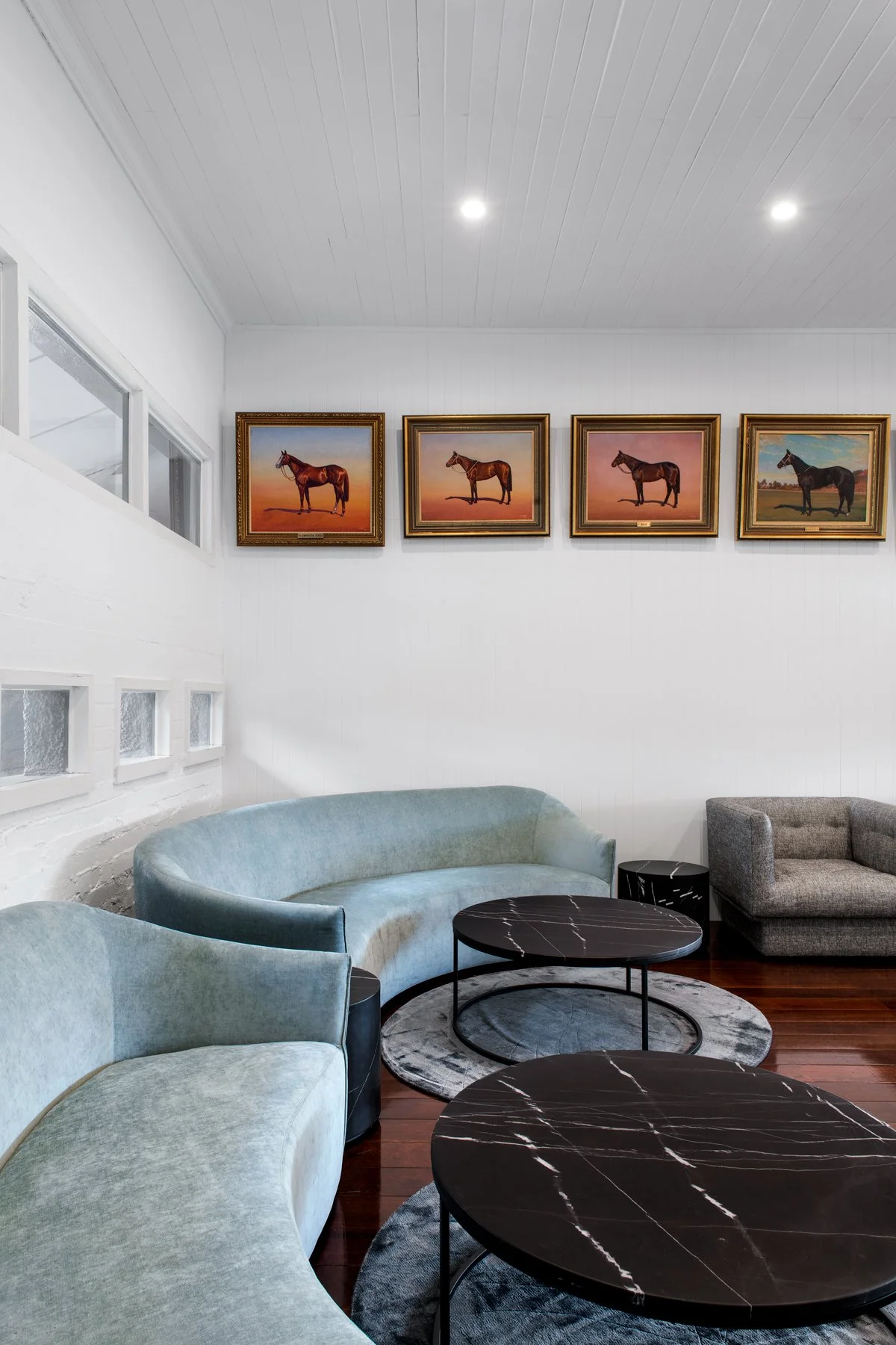 A modern living room with curved sofas in light blue and gray, black marble-top coffee tables, and four framed horse paintings on the white wall.