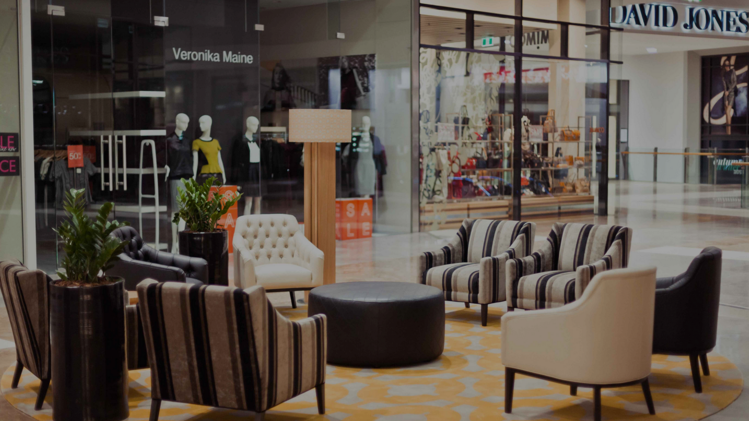 Indoor shopping mall lounge with six chairs, a black ottoman, potted plants, and a floor lamp, with store storefronts and sale signs visible outside.