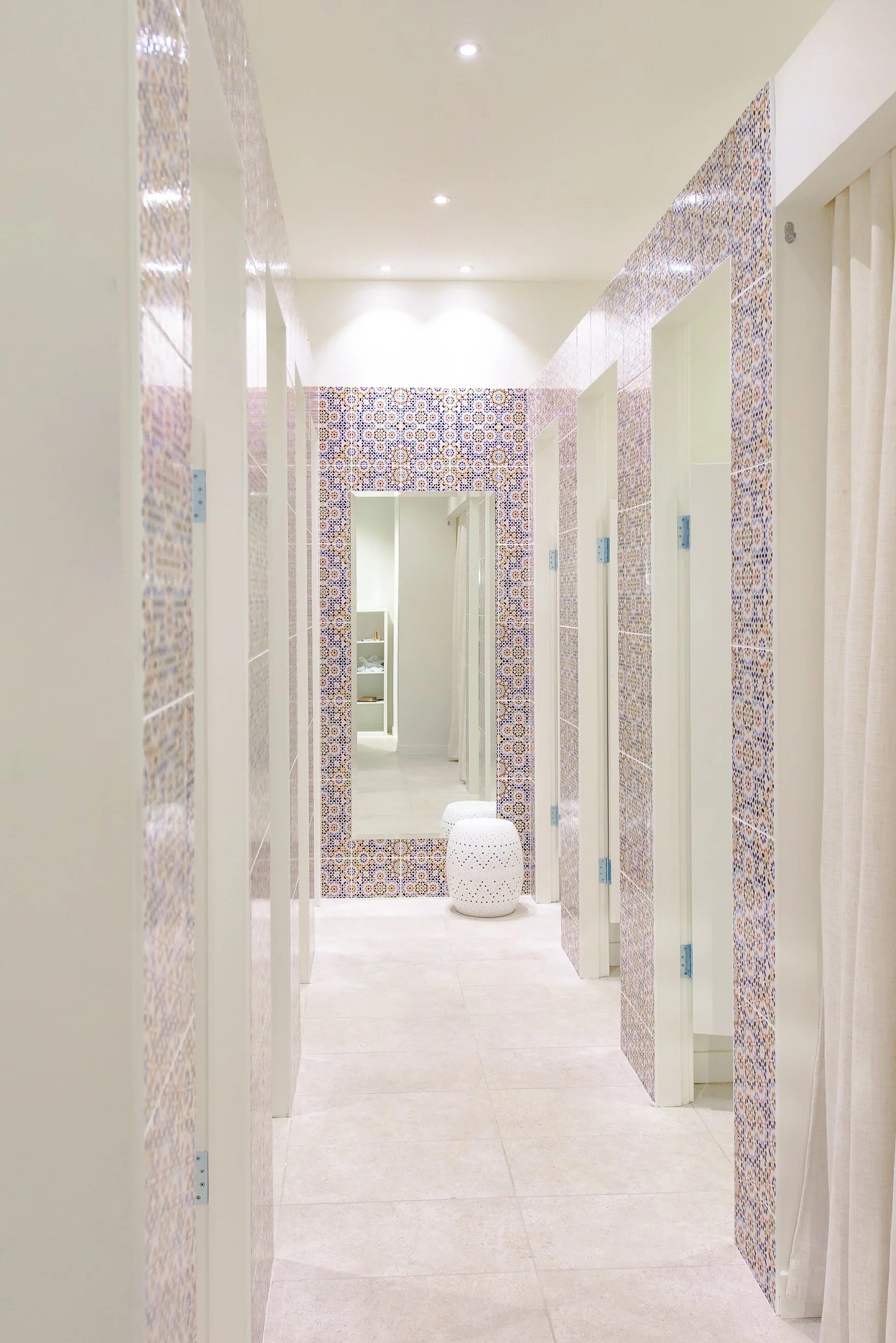 A corridor with multiple bathroom stalls, decorated with geometric patterned tiles on the walls, a large mirror, and a white stool at the end.