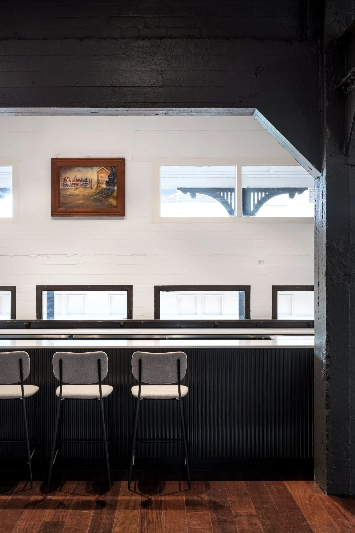 Interior view of a modern space with a black bar-like counter, three gray chairs, a white brick wall, a framed painting, and windows showing an outdoor structure.