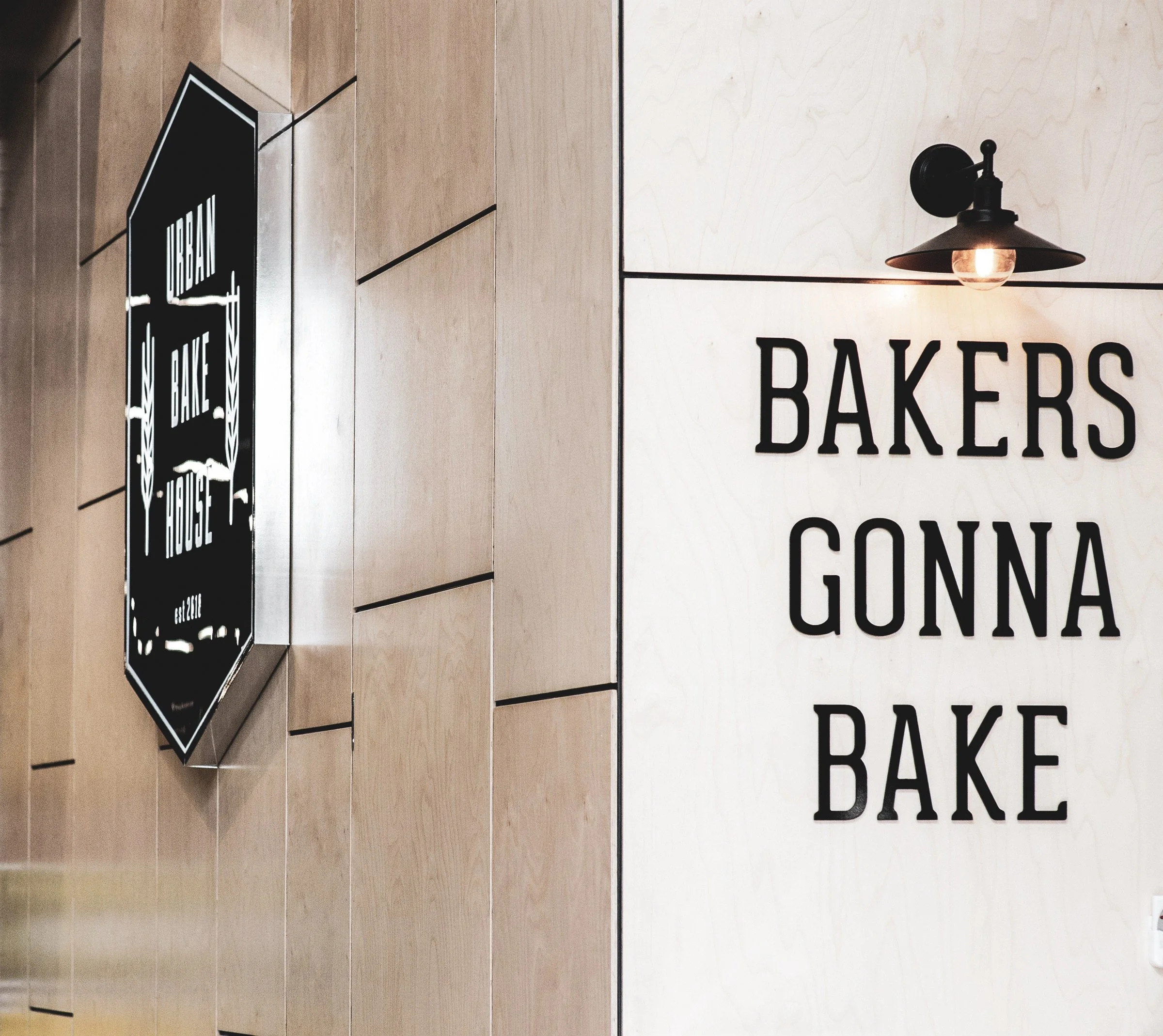 Wall with a black sign that reads 'Urban Bake House' and a white wall with a black lamp and large black text saying 'BAKERS GONNA BAKE'.