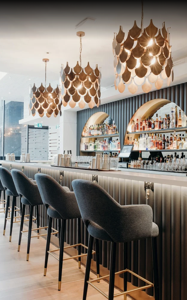 Modern bar interior with high stools, decorative hanging pendant lights, and illuminated display shelves of liquor bottles.