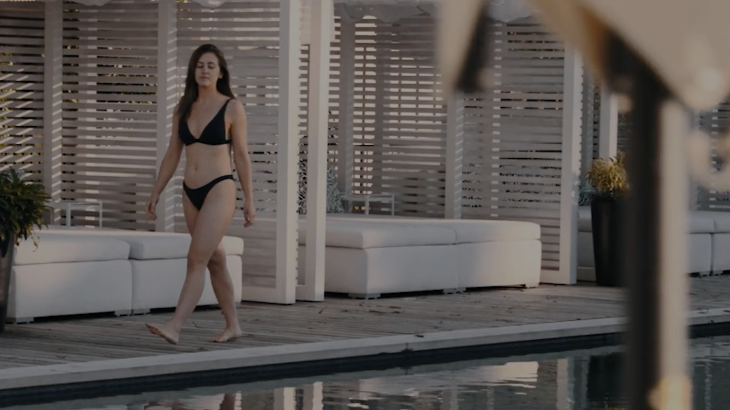 A woman in a black bikini walking along a poolside with modern outdoor furniture and wooden privacy screens in the background.