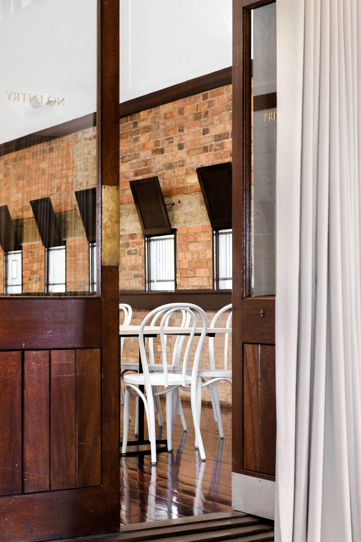 A view through a partially open door revealing white chairs around a table inside a brick-walled room with windows and black wall-mounted objects.