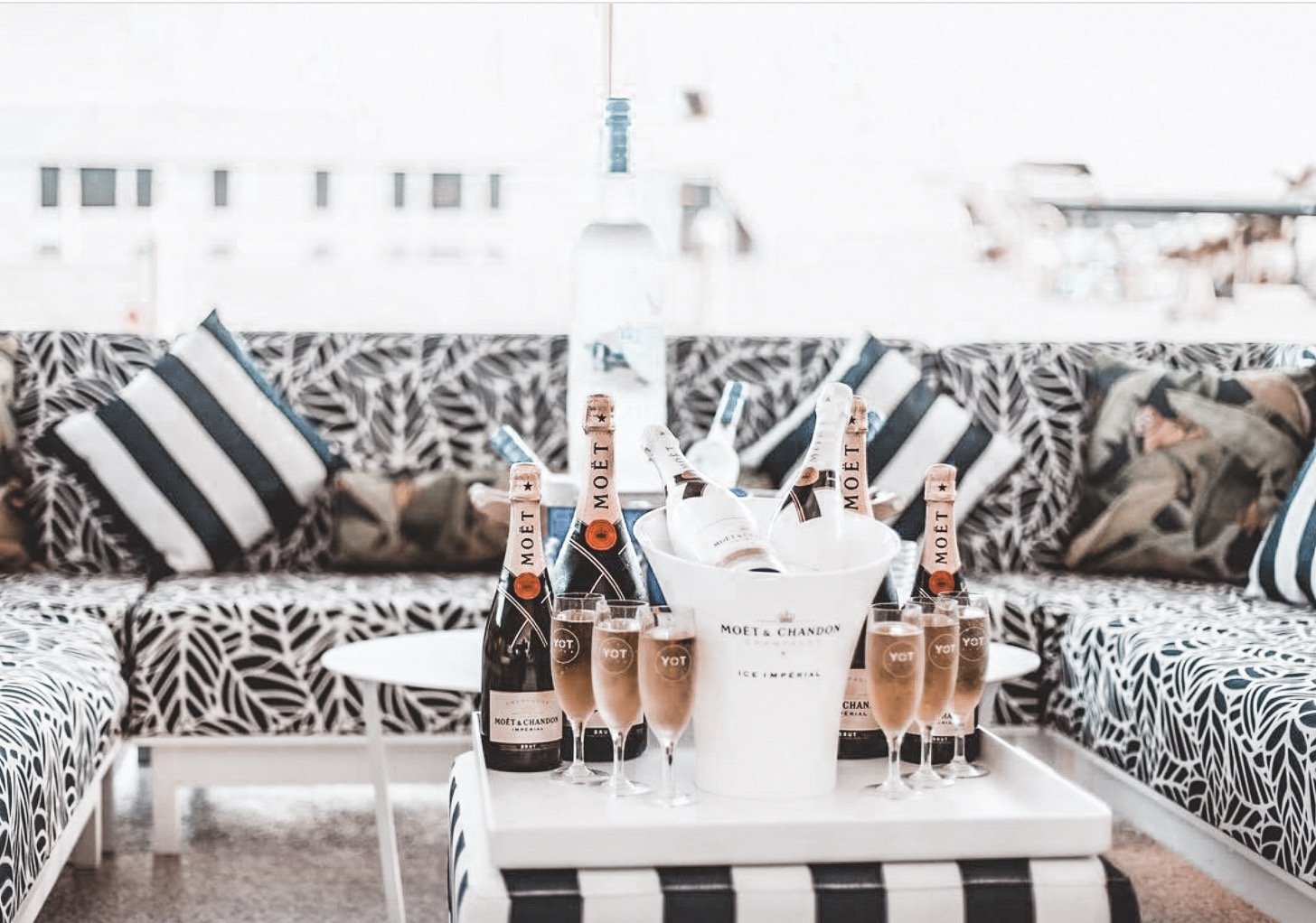A table with several bottles of Moet champagne, champagne glasses filled with champagne, and a white bucket with more bottles inside, set in an outdoor seating area with patterned black-and-white cushions and pillows.