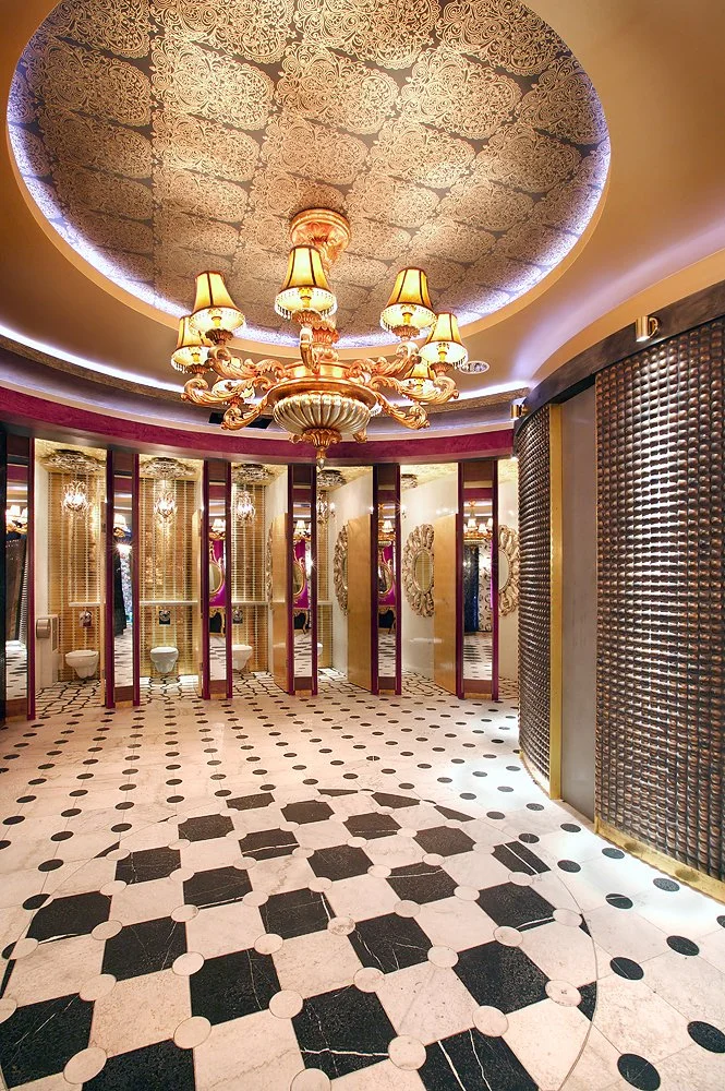 Elegant bathroom with ornate chandelier, decorative ceiling, and individual toilet stalls with mirrors and decorative mirrors inside.
