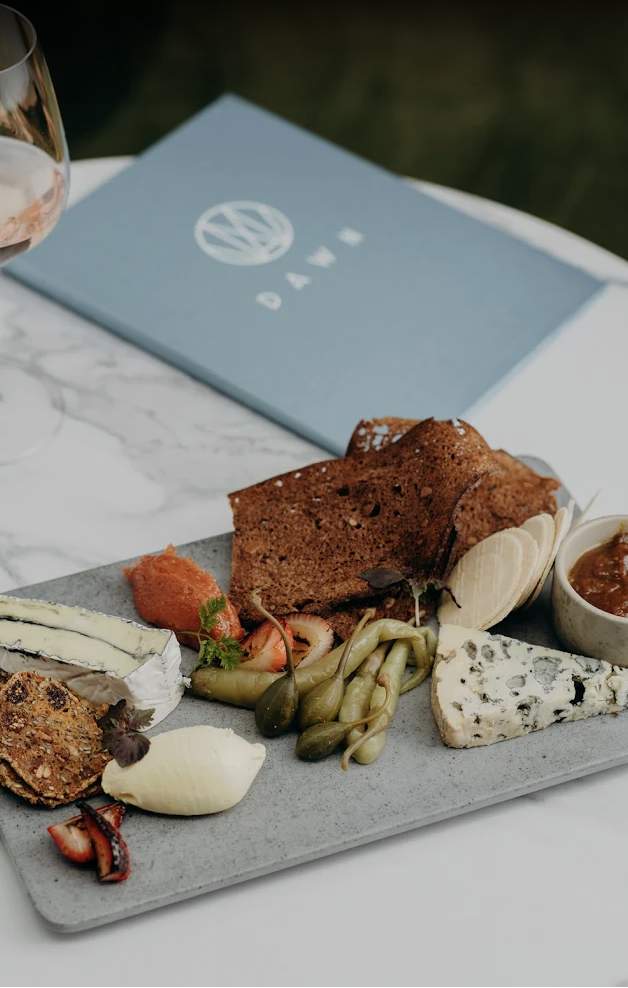 A cheese platter with various cheeses, bread, pickles, strawberries, and a side of jam, on a gray rectangular plate.
