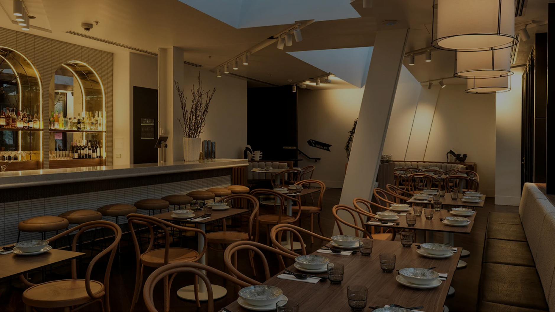 Modern restaurant with wooden tables set with plates, bowls, and glasses, surrounded by wooden chairs and a beige bench seating, with a bar area in the background featuring bottles illuminated under arched glass shelves.