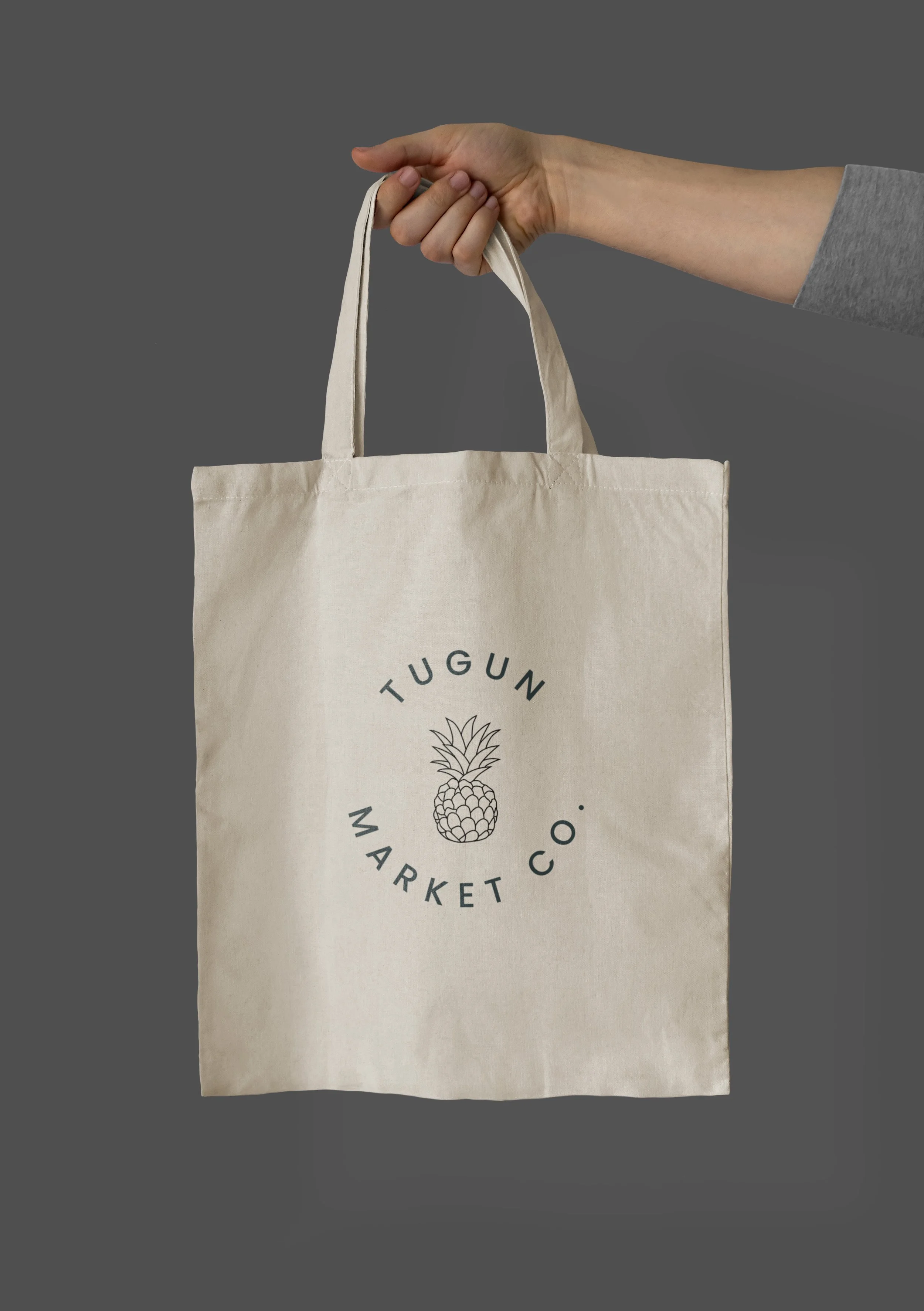A person is holding a beige tote bag with a pineapple graphic and the words 'TUGUN MARKET CO.' printed on it, against a plain gray background.