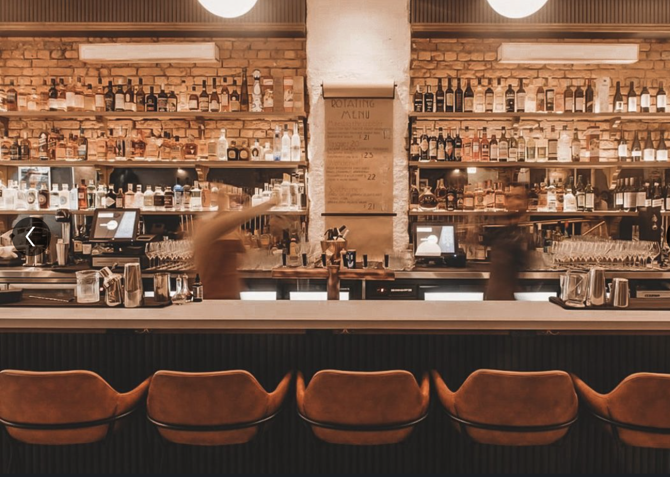 A bar with a brick wall background, shelves stocked with various bottles, a rotating menu sign, and four brown bar stools in the foreground.