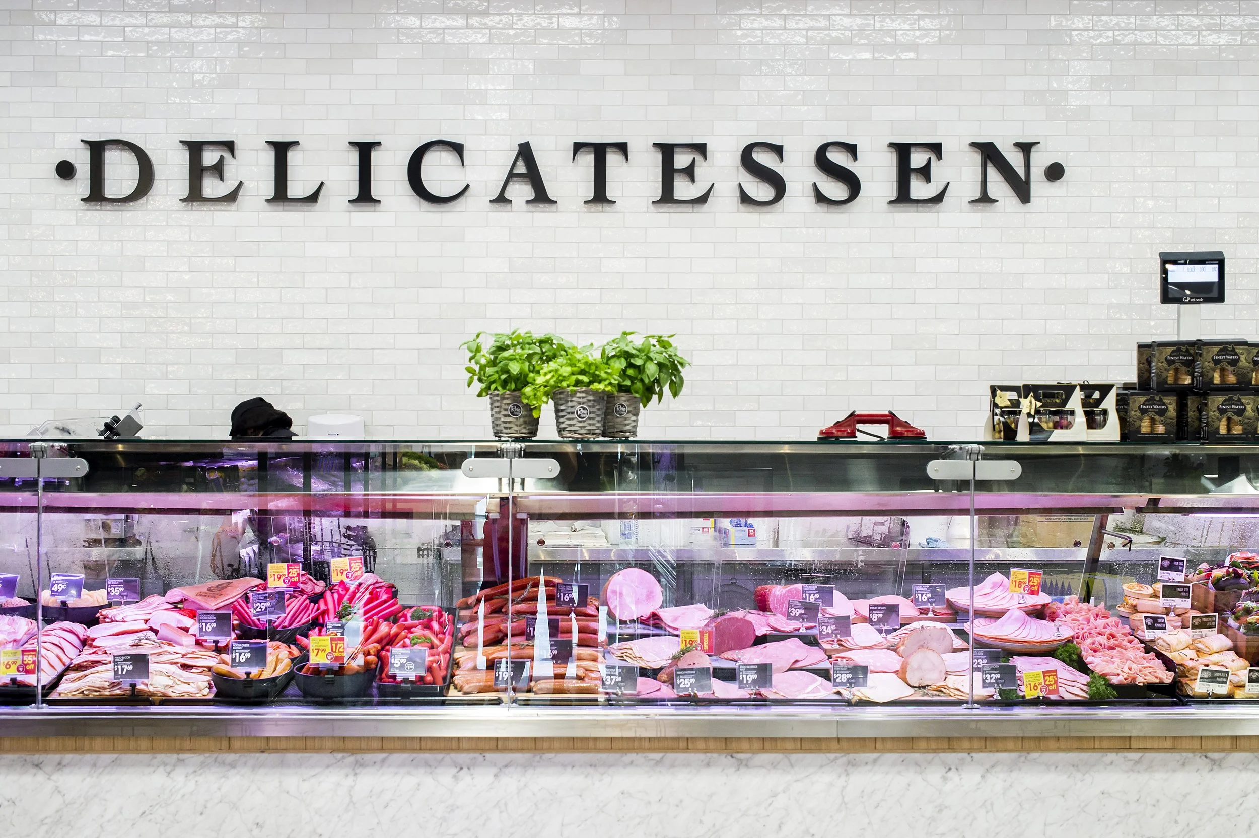 A deli counter with various meats and sausages in a glass display case, a sign above reading 'Delicatessen', and plants on top of the counter.