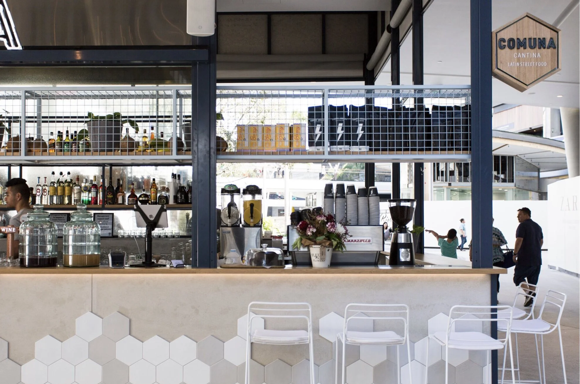 Modern cafe counter with coffee machines, a flower arrangement, and upscale drink dispensers in a well-lit, open space with white hexagonal tile design on the front.