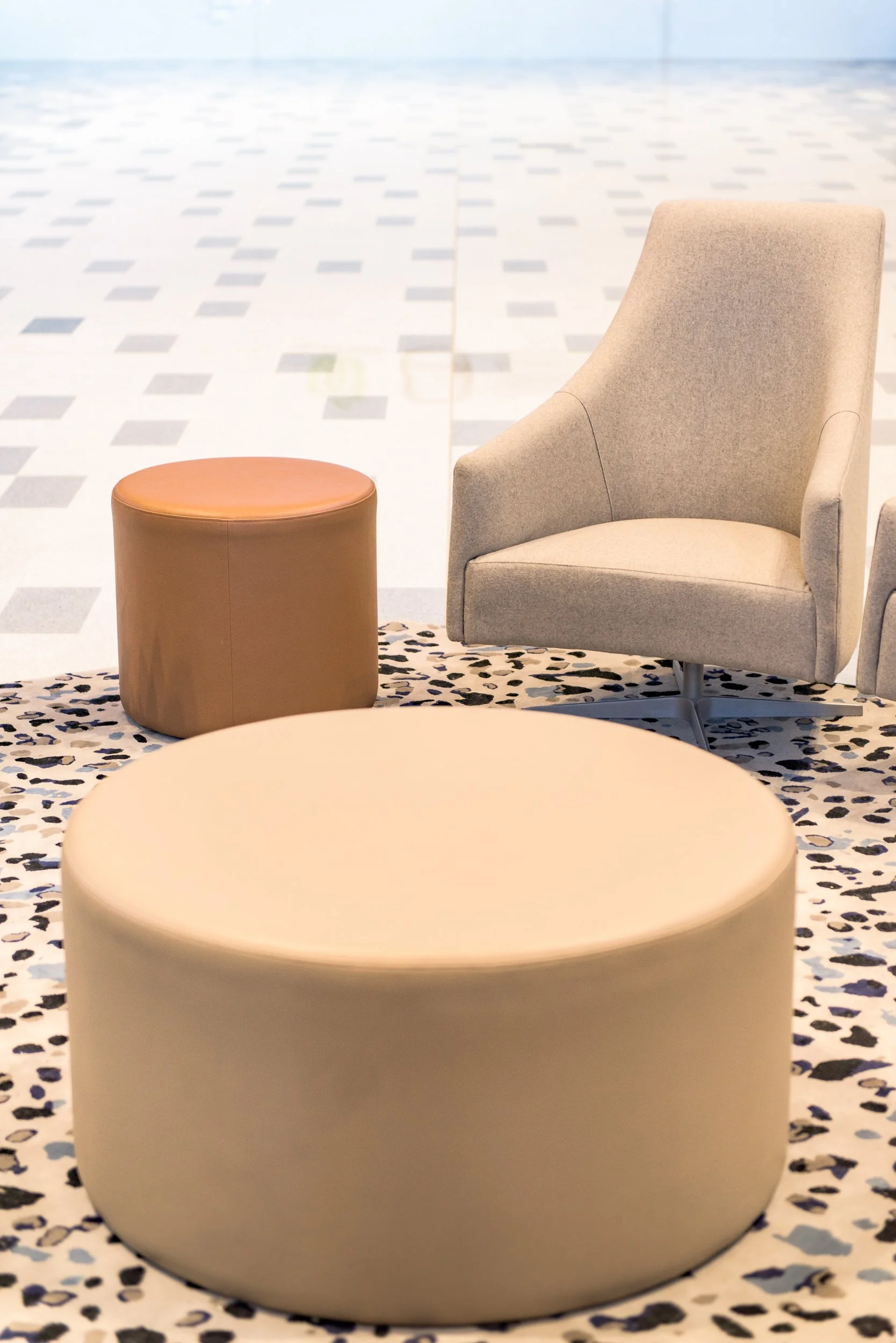 Modern seating area with a beige armchair, a beige ottoman, and a round beige coffee table on a patterned rug, against a background of an open, empty space with tiled floor.