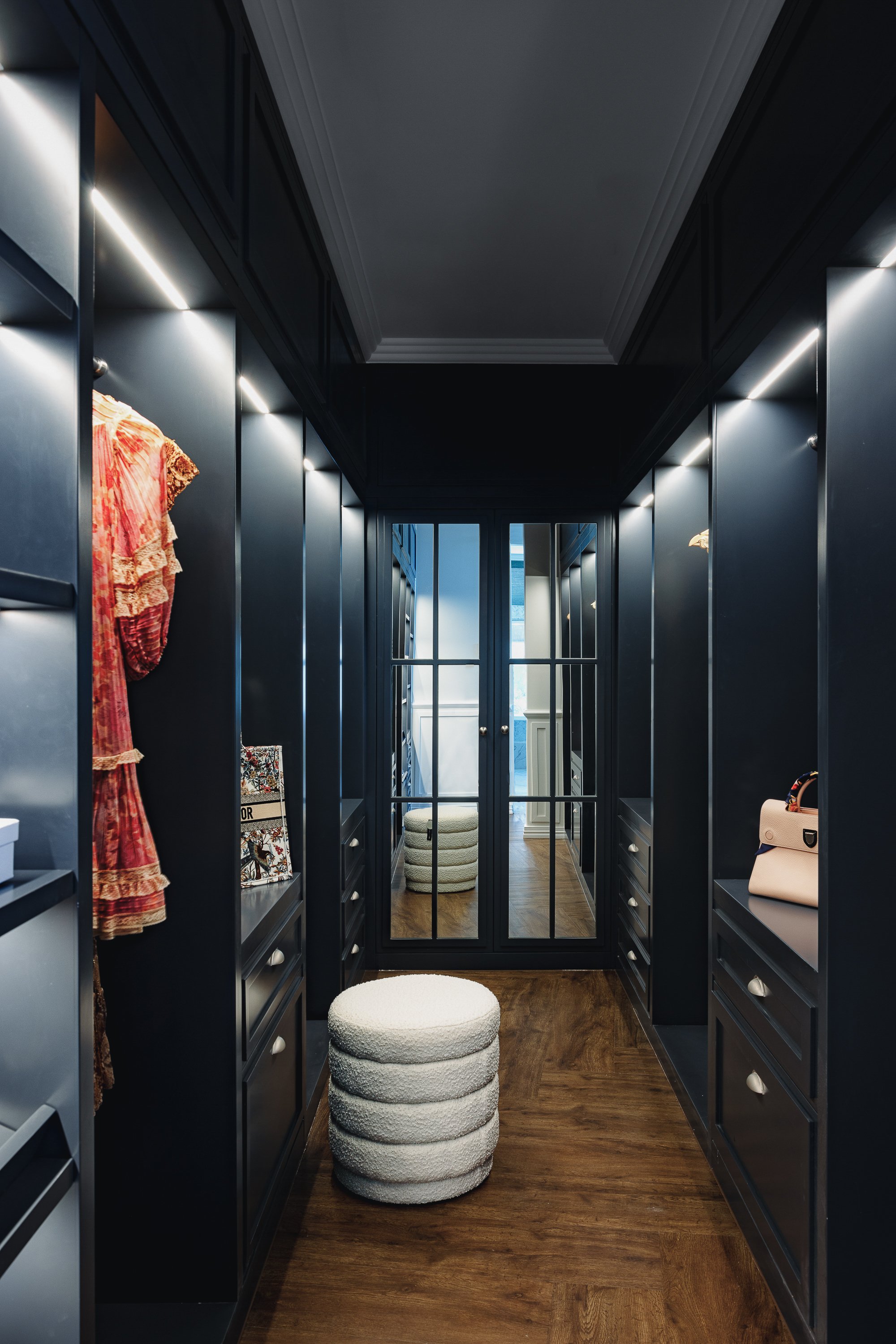 Elegant walk-in closet with dark cabinetry, wooden floor, and LED strip lighting. Features a white textured stool, mirrored doors, a pink handbag, and a rack with colorful clothing.