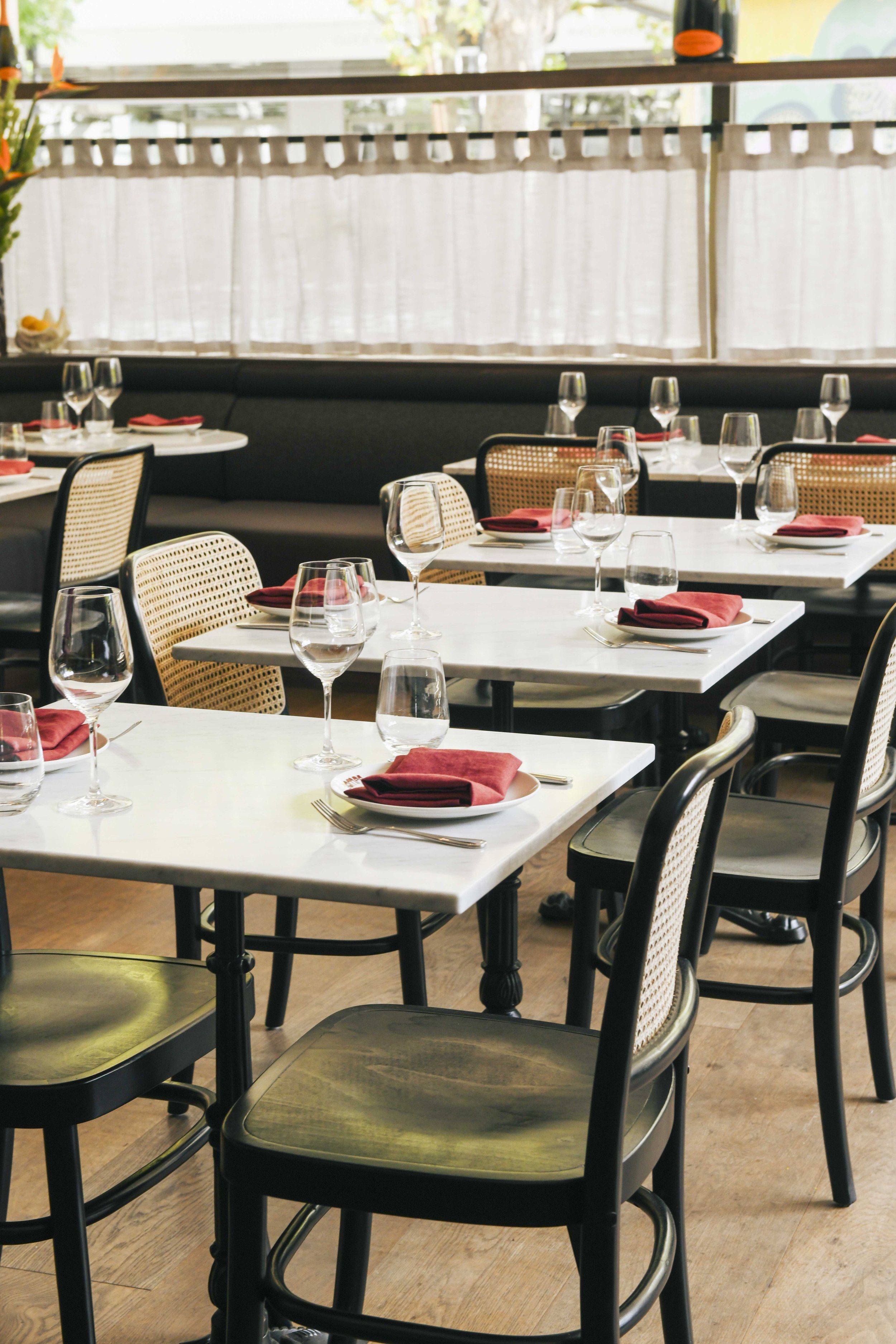 Interior of a restaurant with white marble tables, black chairs with cane backs, arranged place settings, and red napkins.