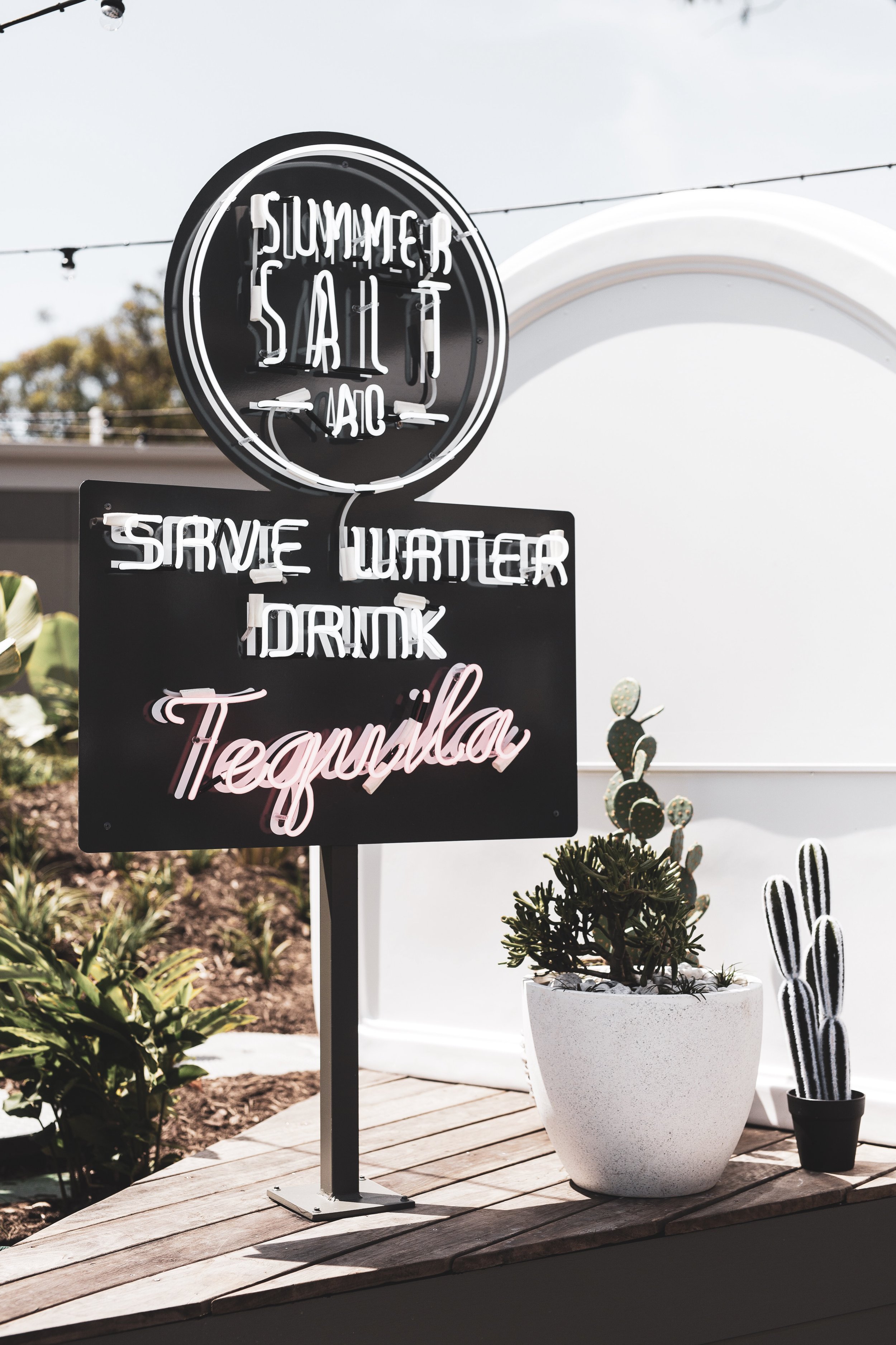 A neon sign outside a restaurant or bar advertising summer patio food and drinks, including tequila, with a potted cactus plant nearby.