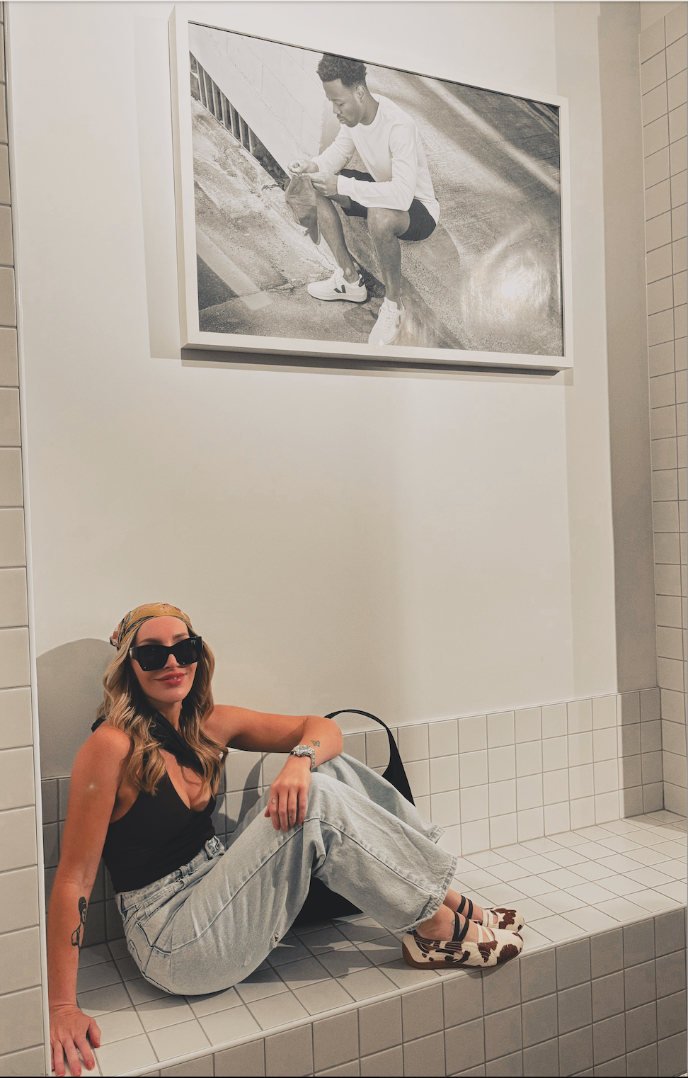 A woman with blonde hair, wearing large black sunglasses, a patterned headscarf, a black tank top, light gray baggy jeans, and patterned shoes, sitting on a tiled bench in an art gallery. Behind her, a large framed black-and-white photo of a man squatting by a curb, wearing a white long-sleeve shirt, dark shorts, and sneakers.