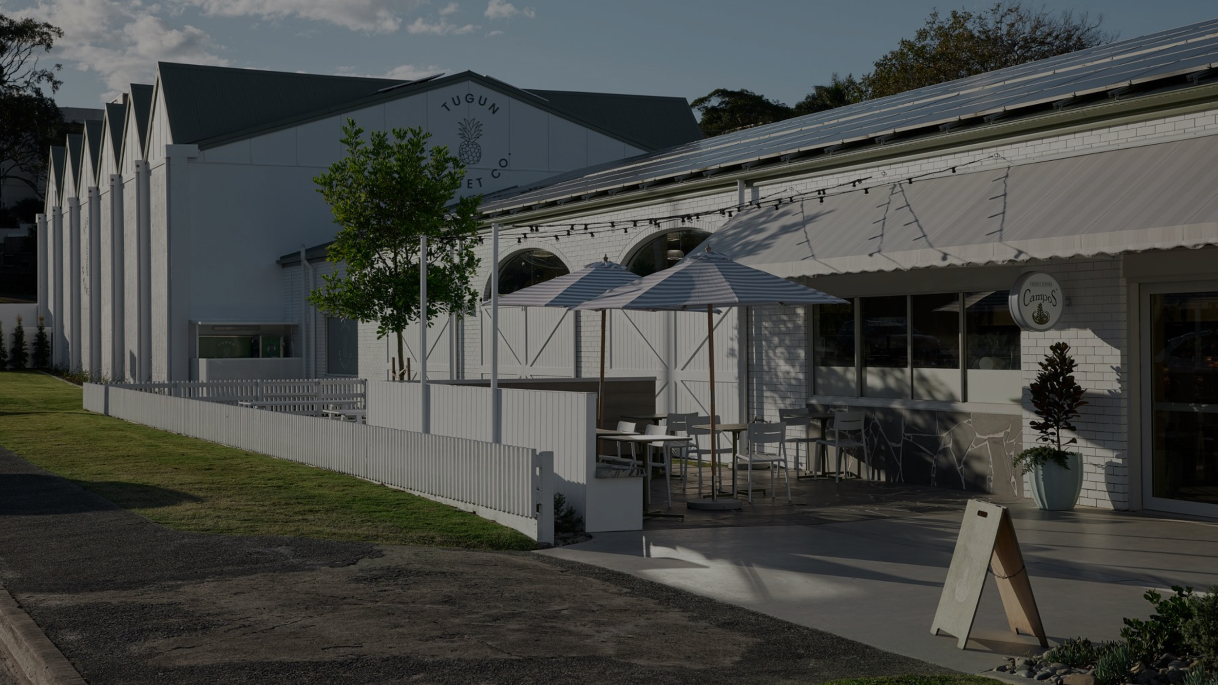 Exterior view of a white building with outdoor seating shaded by umbrellas, potted plants, and a sign reading 'Campos.'