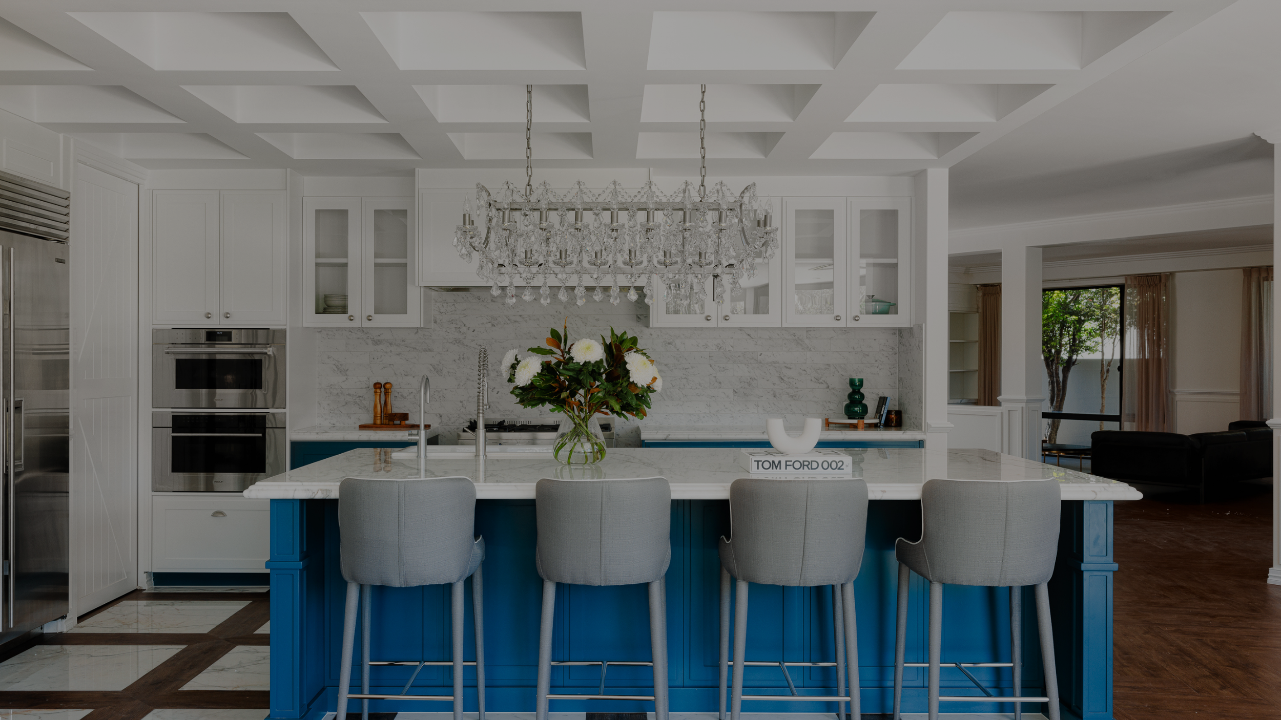 Modern kitchen with a white marble island, blue base, gray chairs, crystal chandelier, white cabinetry, and a vase of white flowers.