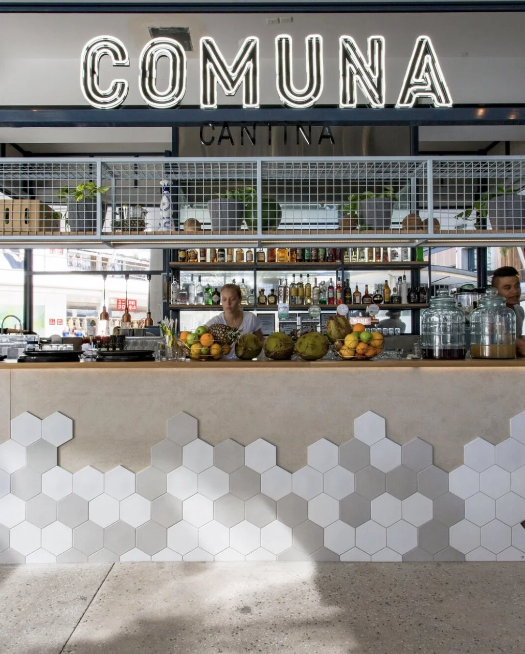 Modern restaurant bar with a backlit sign reading 'COMUNA', a bar counter with fruits like pineapples and coconuts, glass jars, bottles of alcohol, and bartenders preparing drinks. White hexagon tiles decorate the front of the bar.