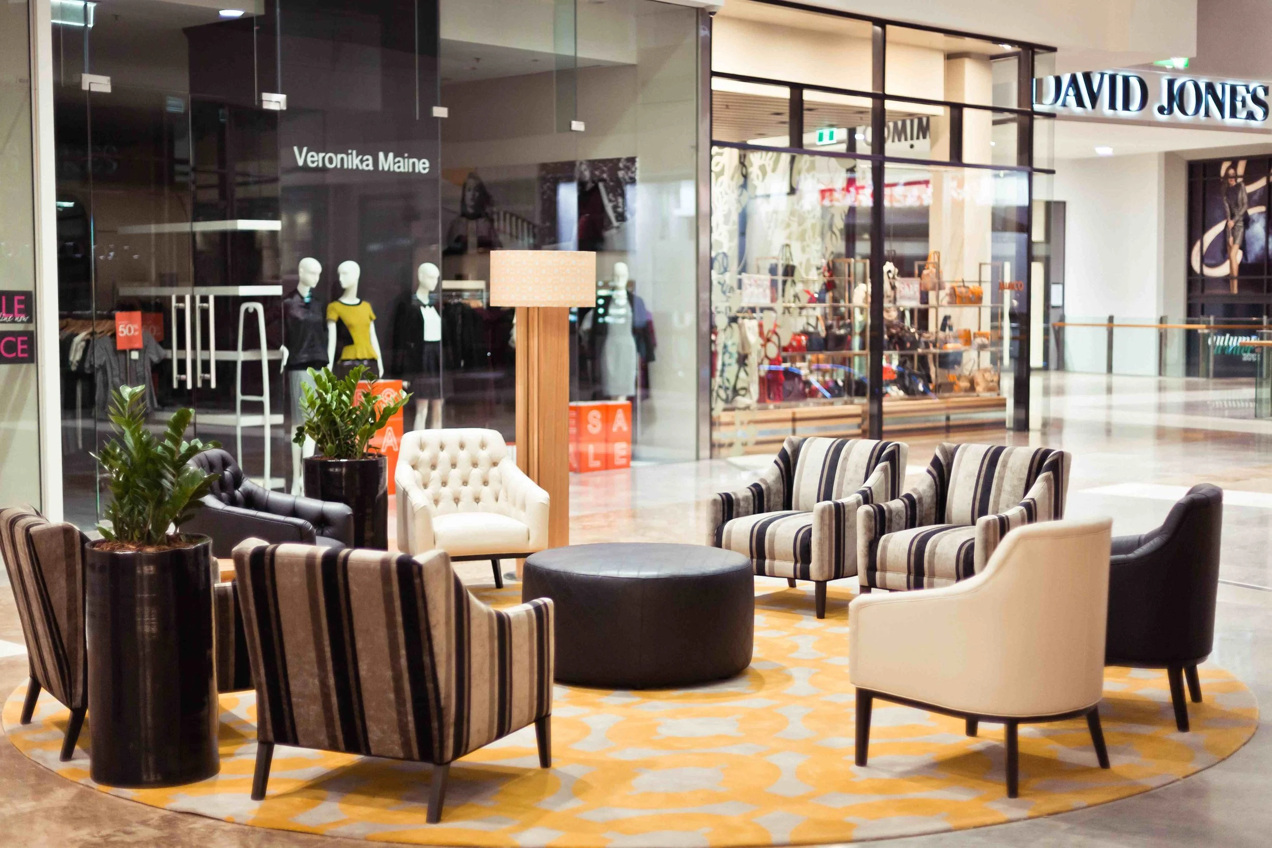 A seating area inside a shopping mall featuring a round yellow and gray rug, various chairs including striped, black, cream, and tufted styles, a black ottoman, a wooden lamp with a beige shade, and two large potted plants. In the background, storefr