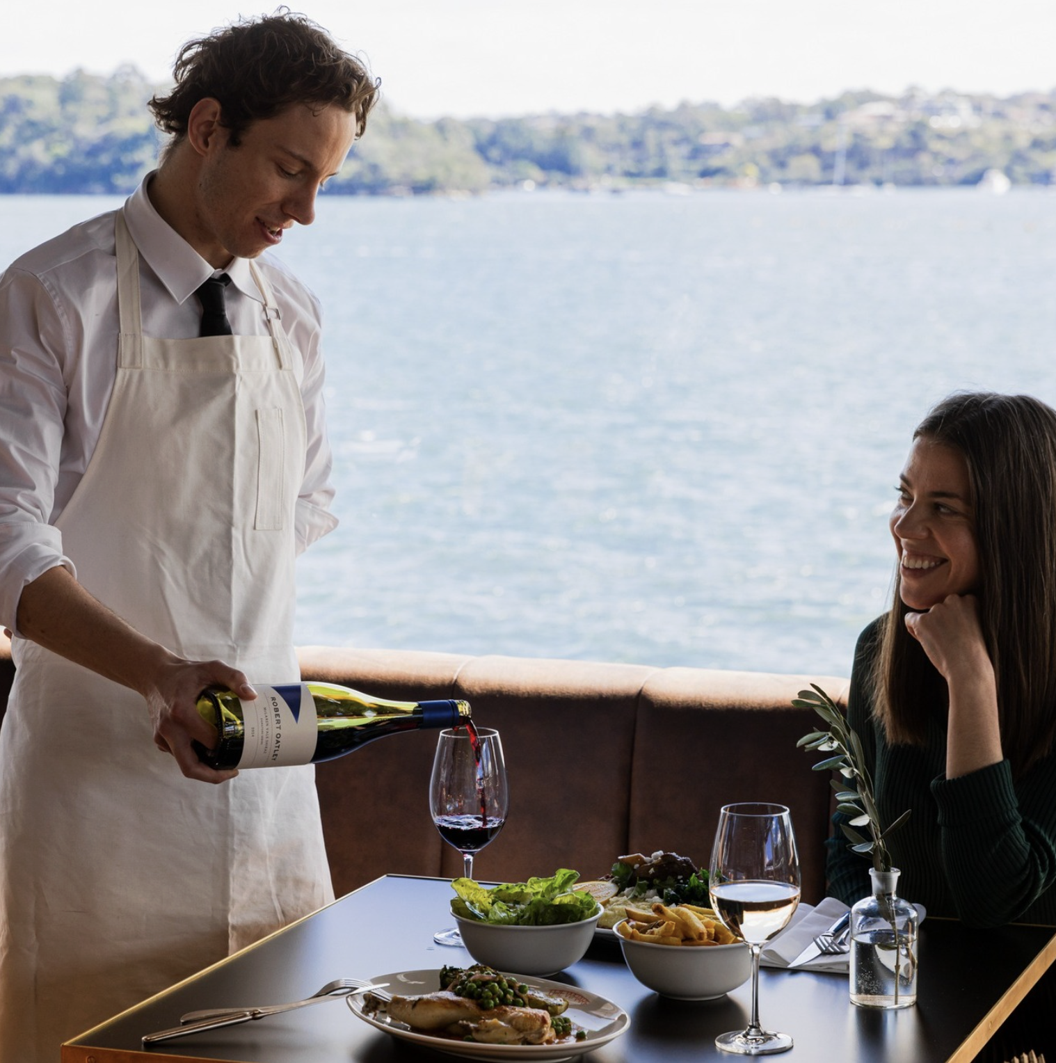 A waiter pouring red wine into a glass for a woman at a seaside restaurant. The woman is smiling and sitting at a table with salads, a plate of food, and drinks. The background shows water and distant shoreline.